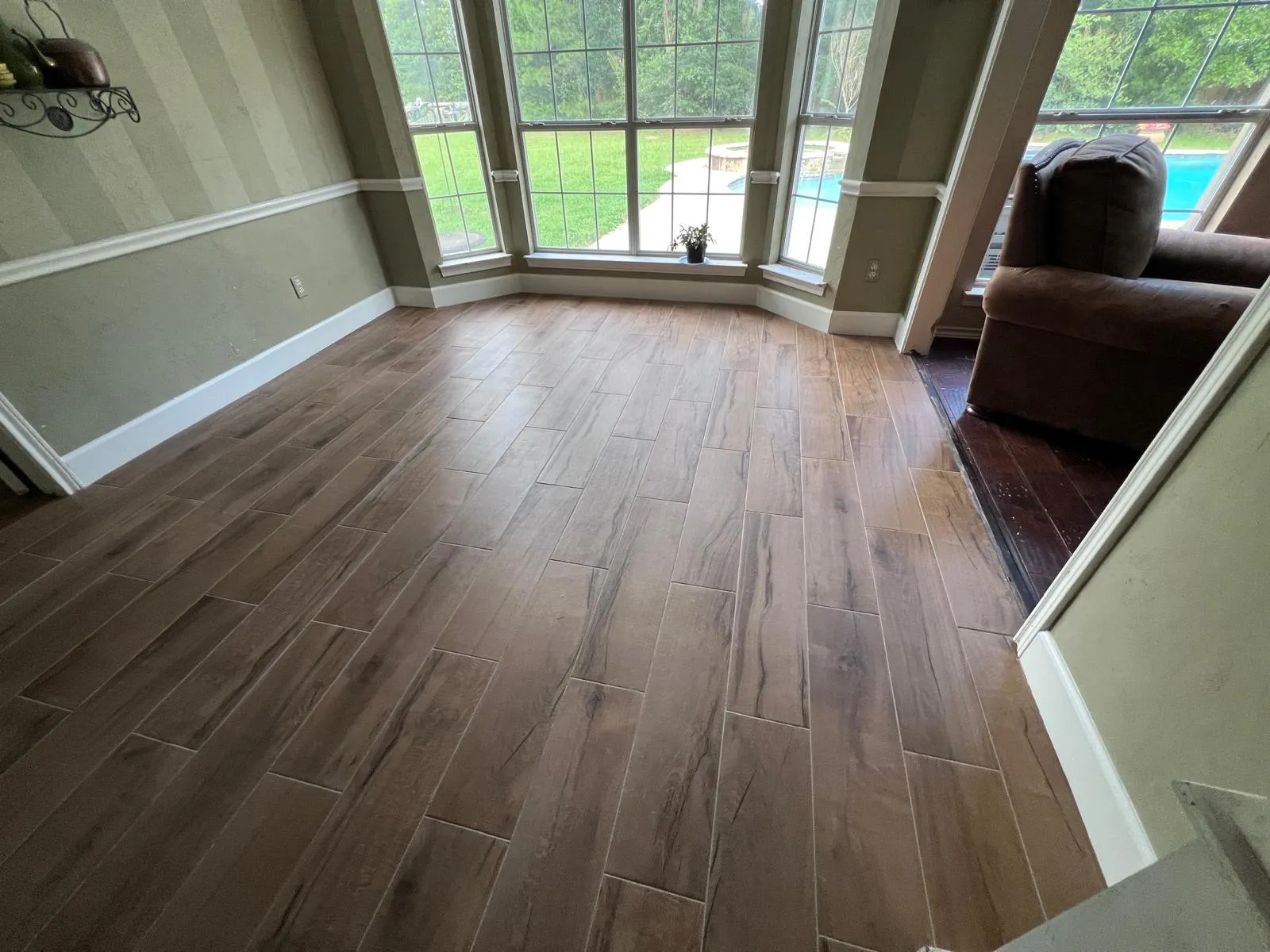 Bright room with wood-look tile flooring, large windows overlooking a green lawn and pool, and a brown upholstered chair.
