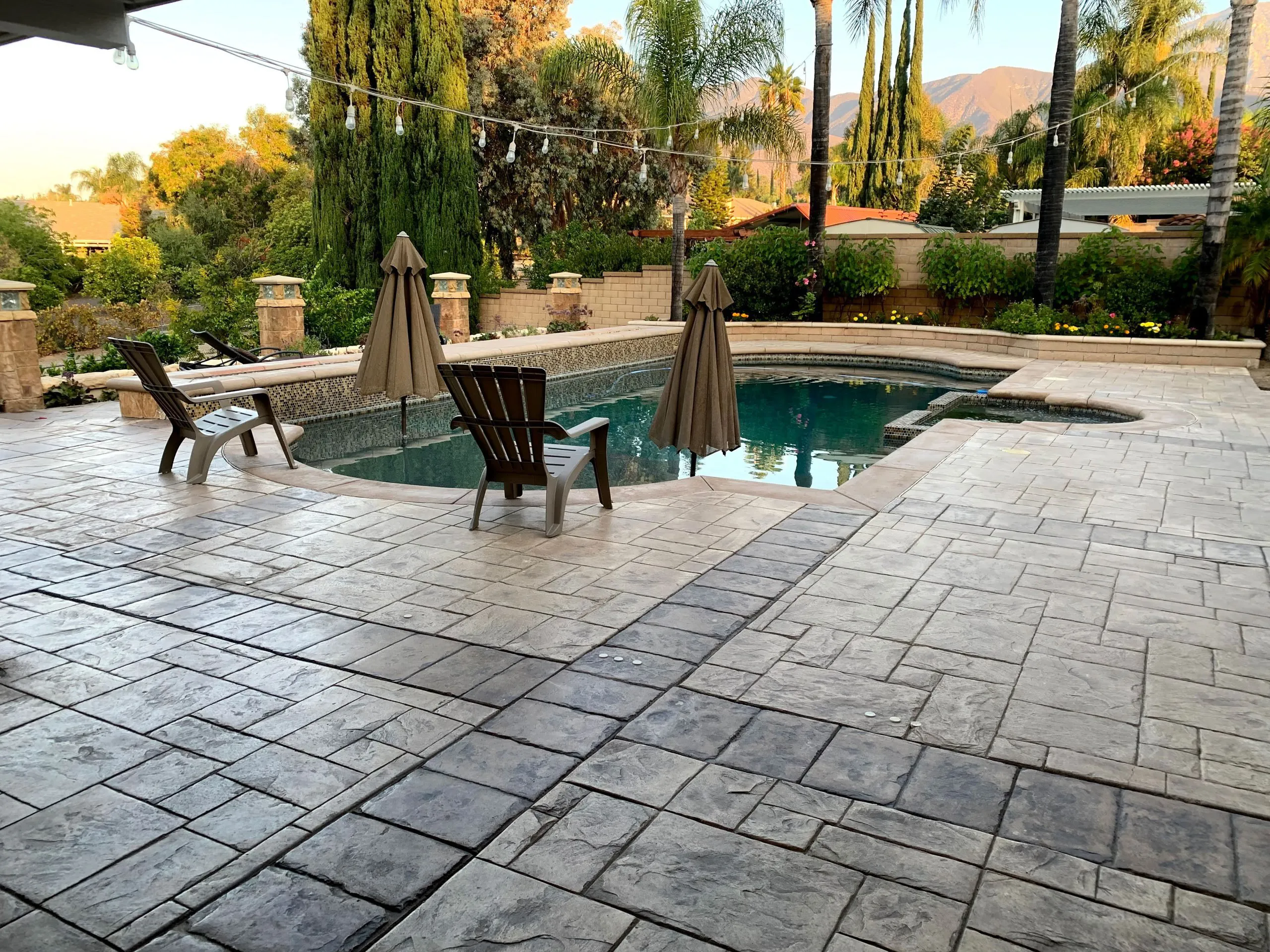 Backyard swimming pool surrounded by stamped concrete patio, two brown umbrellas, and two plastic Adirondack chairs, with tall trees and mountains in the background.