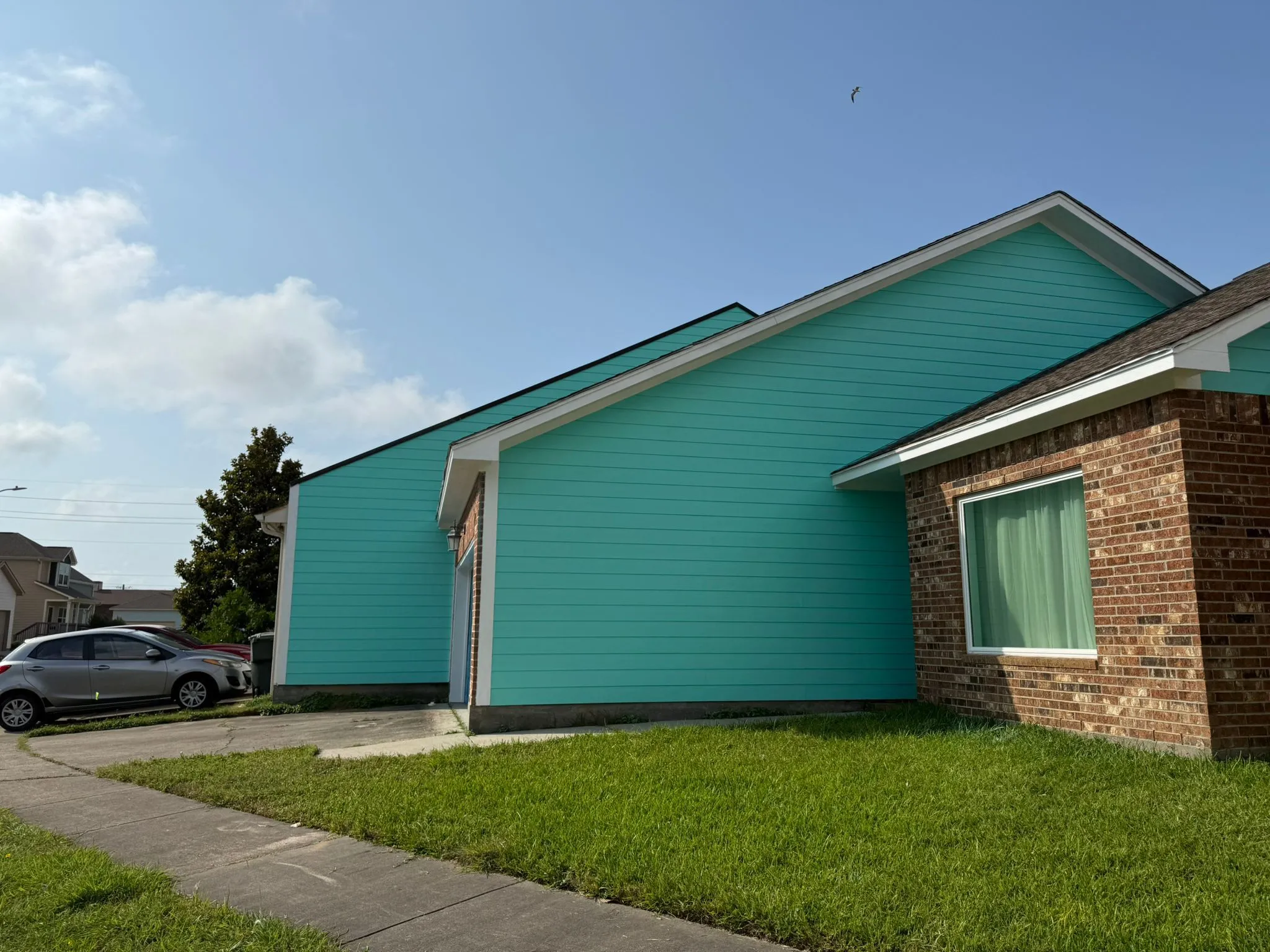 Side view of a turquoise house with brick accents, green lawn, and a car parked on the driveway under a partly cloudy sky.