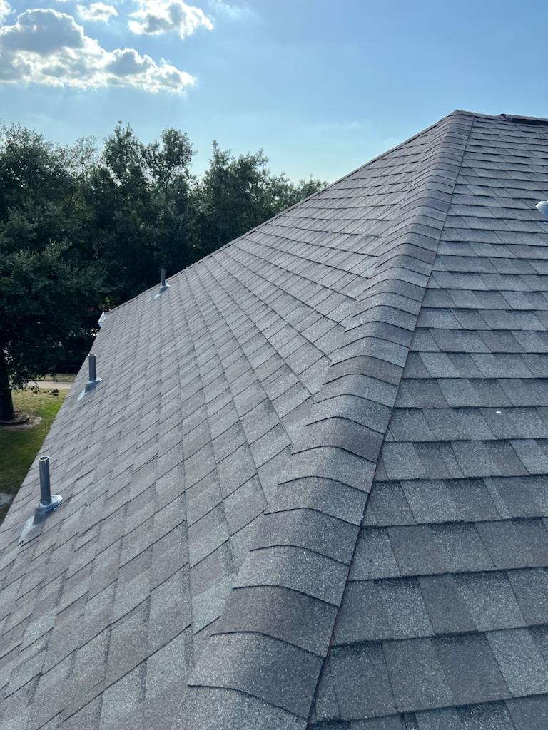 Gray shingled roof with ridge cap under a partly cloudy blue sky and trees in the background.