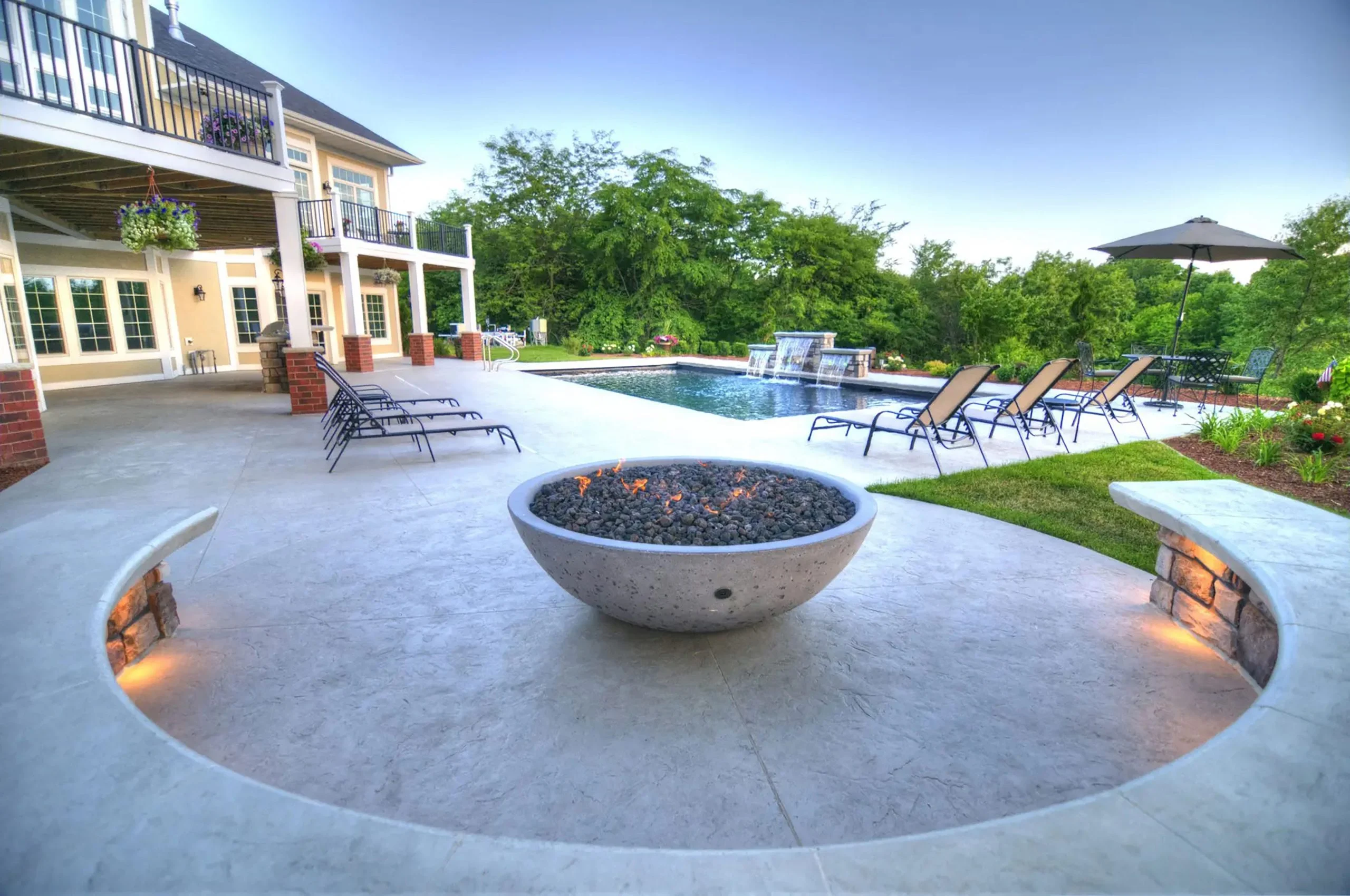 Modern backyard with a curved concrete bench, fire pit, lounge chairs, swimming pool with waterfalls, and patio umbrella.