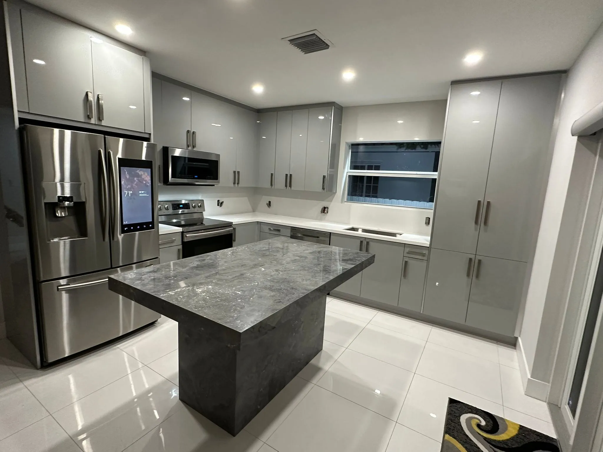 Modern kitchen with glossy gray cabinets, stainless steel appliances, and a marble island under recessed ceiling lights.