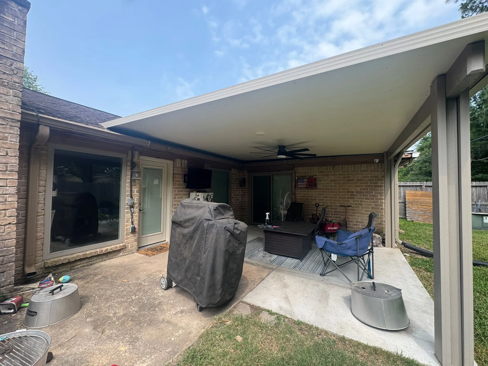 Covered patio with a ceiling fan, a covered grill, outdoor chairs, and various items on a concrete and grass yard.