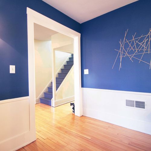 Interior view of a room with blue upper walls, white lower walls, hardwood floor, blue staircase, and a black-and-white cat peeking around a white door frame.
