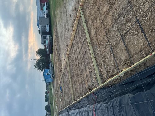 Construction site showing a prepared foundation with wooden frames and metal rebar grid on dirt under a cloudy sky.