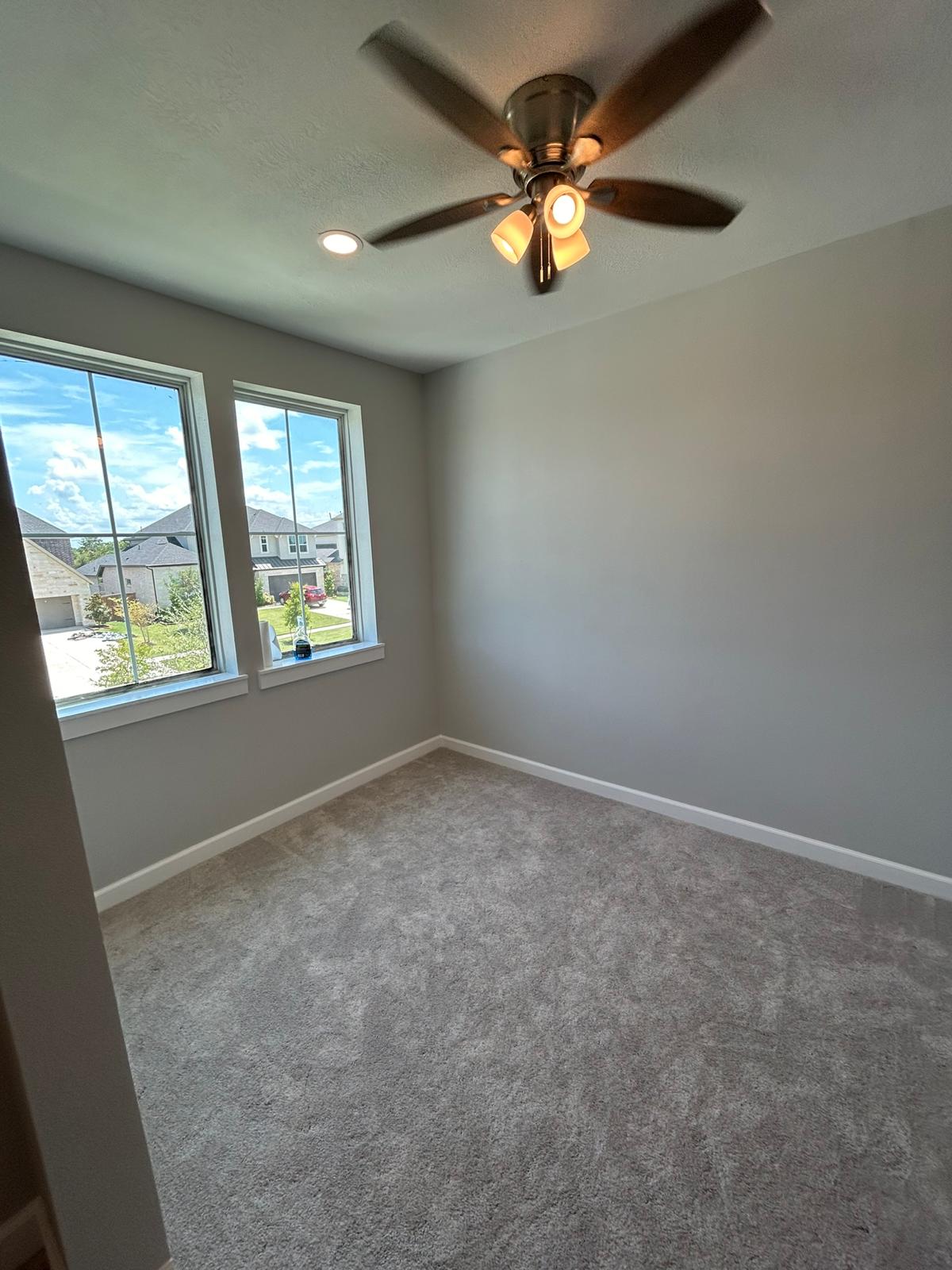 Empty room with light gray walls, carpeted floor, two windows showing suburban houses, and a ceiling fan with lights.