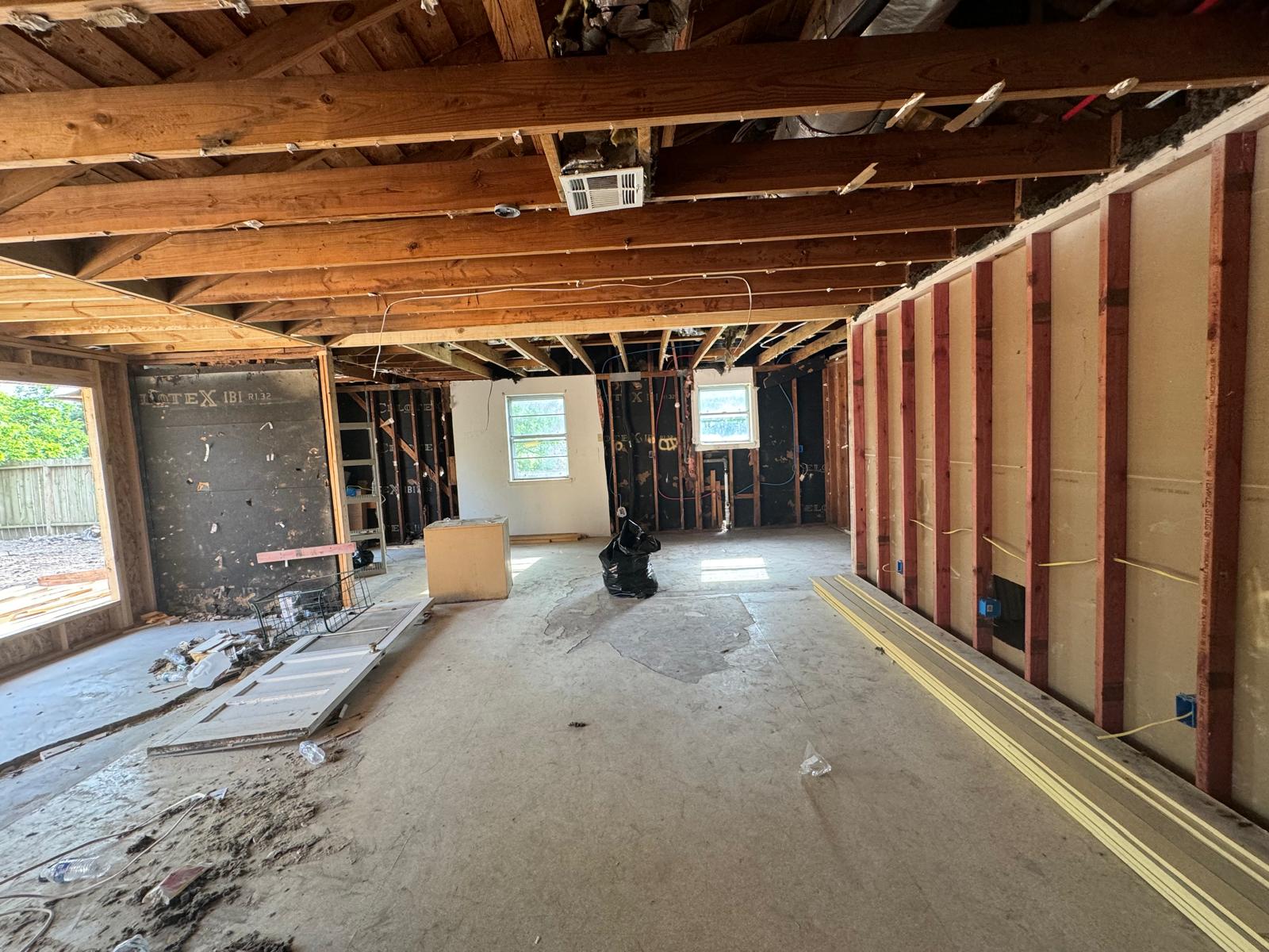 Interior of a residential construction site showing exposed wooden ceiling beams, unfinished walls with electrical wiring, construction materials, and debris on the floor.