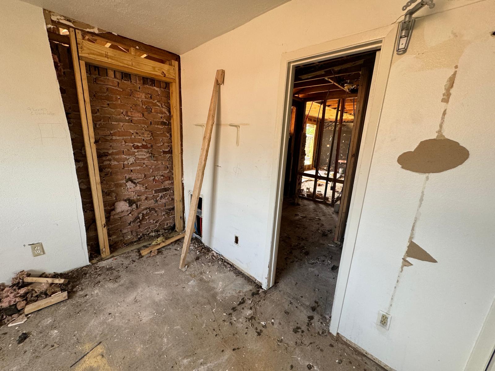 Interior room under renovation with exposed brick wall framed by wood, a wooden plank leaning against a white wall, and a doorway leading to another unfinished room with exposed wooden beams.