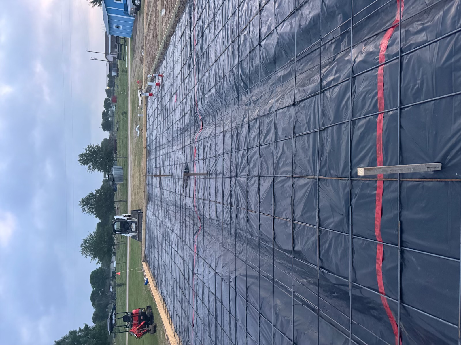 Construction site with foundation prepared with black plastic sheeting and metal rebar grid, small excavators and trees in the background.