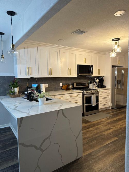 Modern kitchen with white cabinets, gold handles, marble countertop island, stainless steel appliances, wood flooring, and pendant lights.