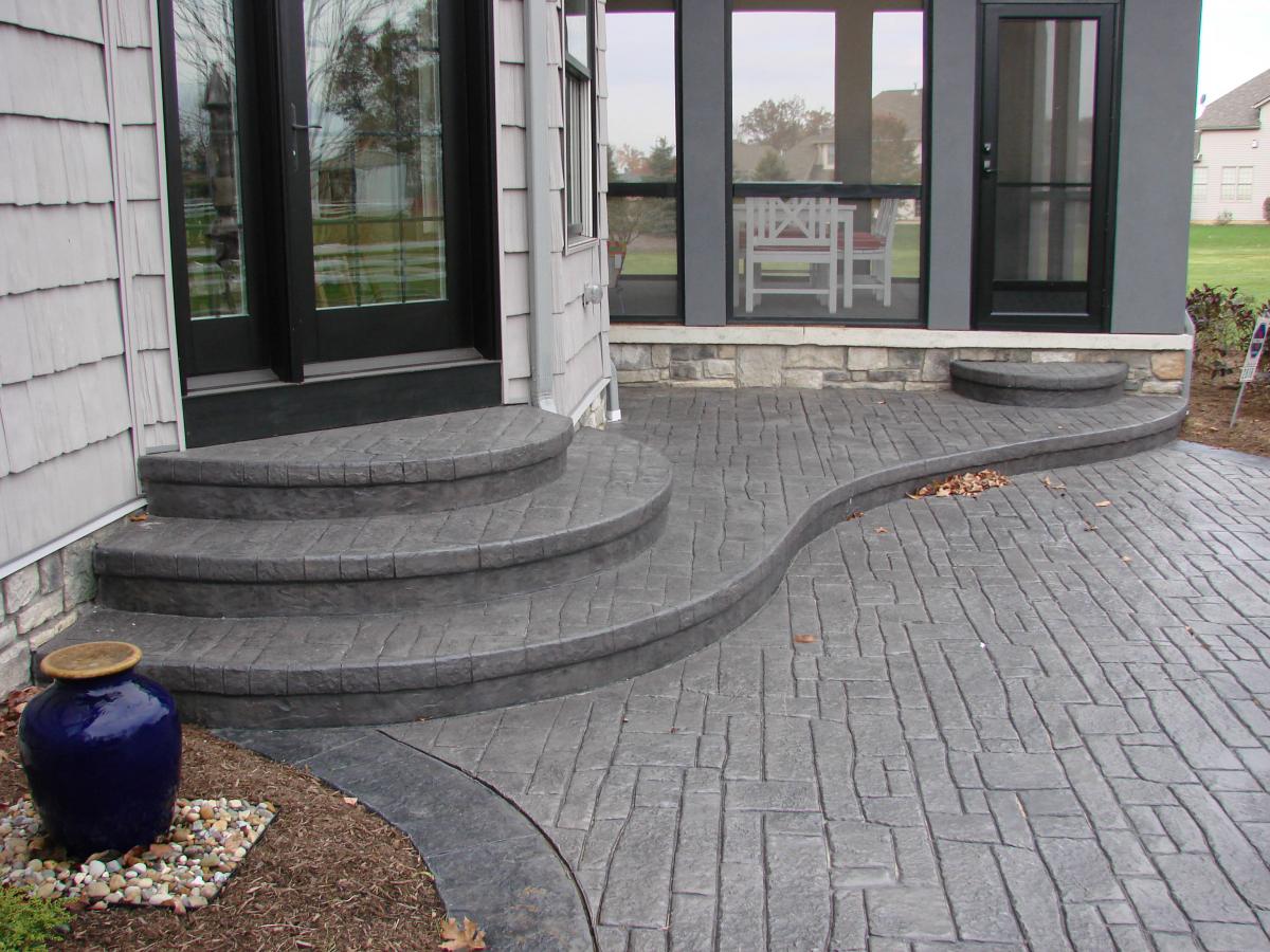 Curved stamped concrete patio with steps leading to a black-framed glass door, a blue decorative urn on a mulched area, and a screened-in porch with white chairs.