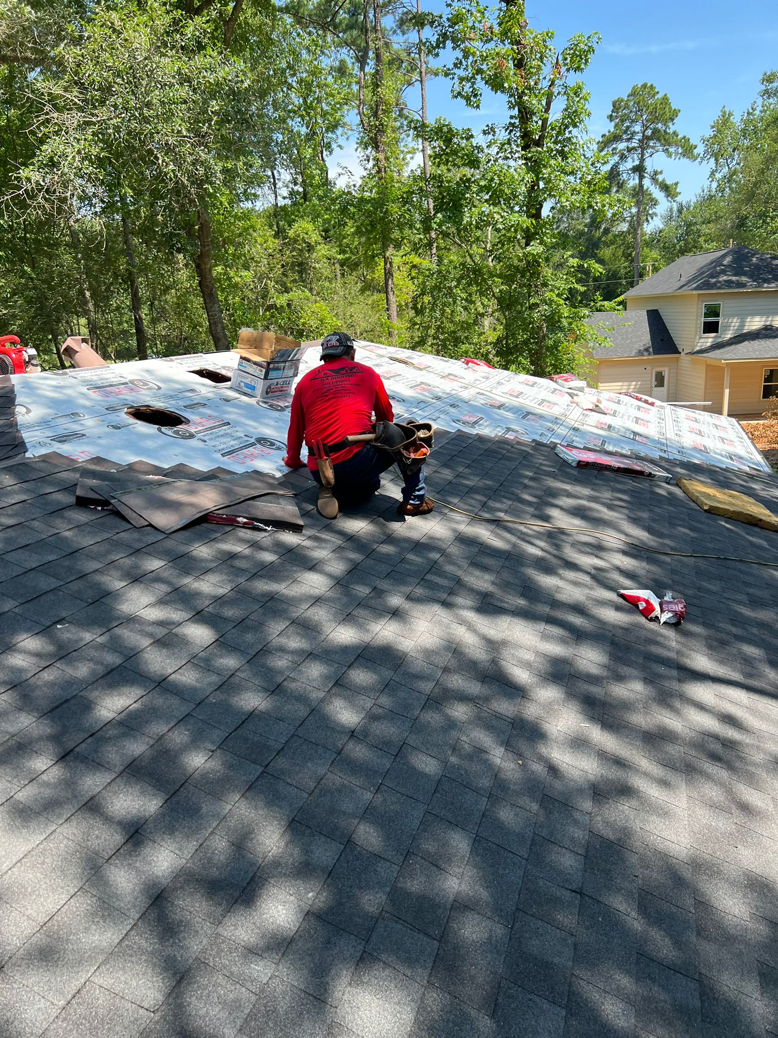 Roofer wearing a red shirt installing shingles on a roof under a clear blue sky with trees and nearby houses in the background.