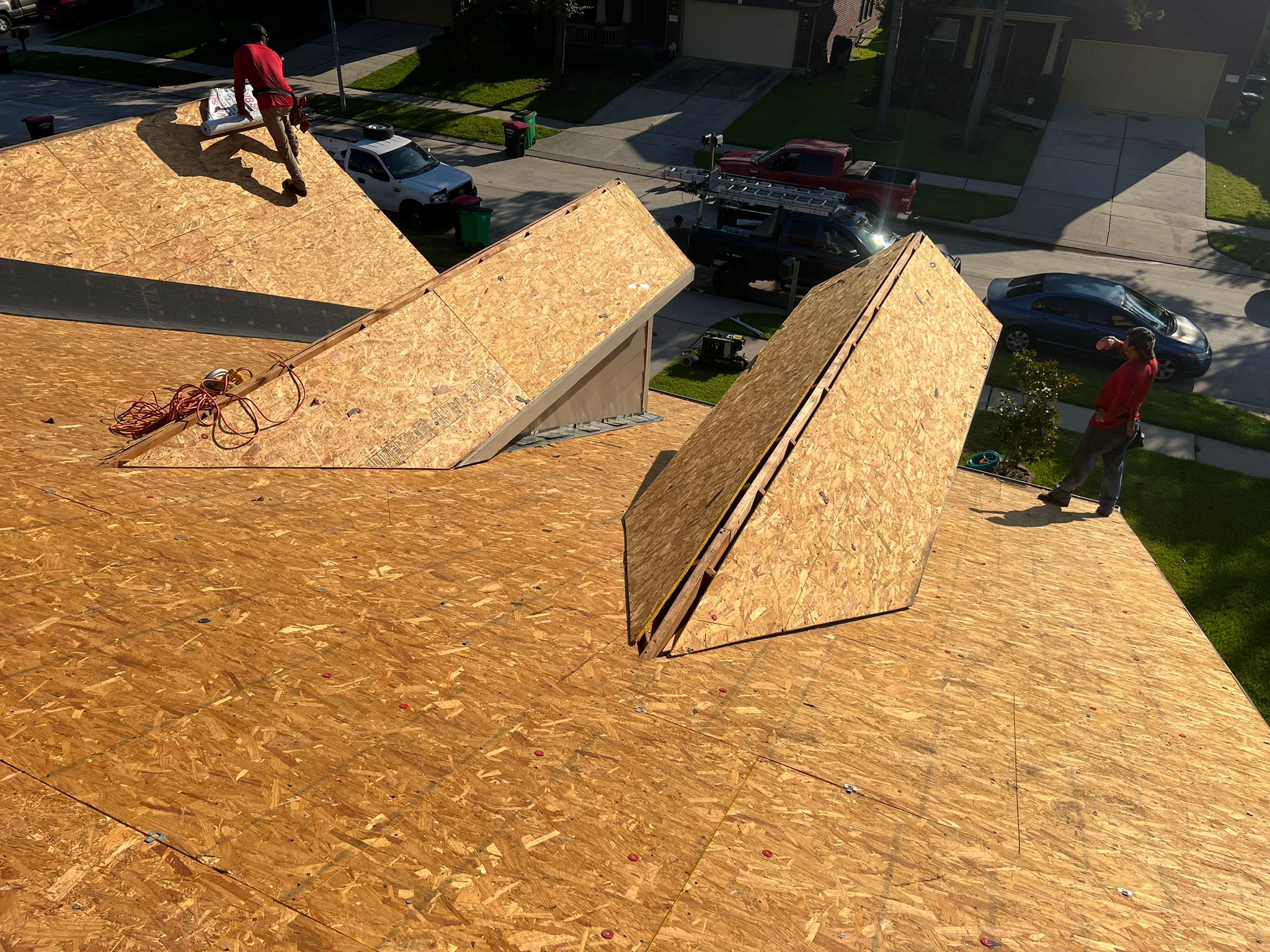 Two workers on a newly plywood-covered roof near two dormer structures under bright sunlight.