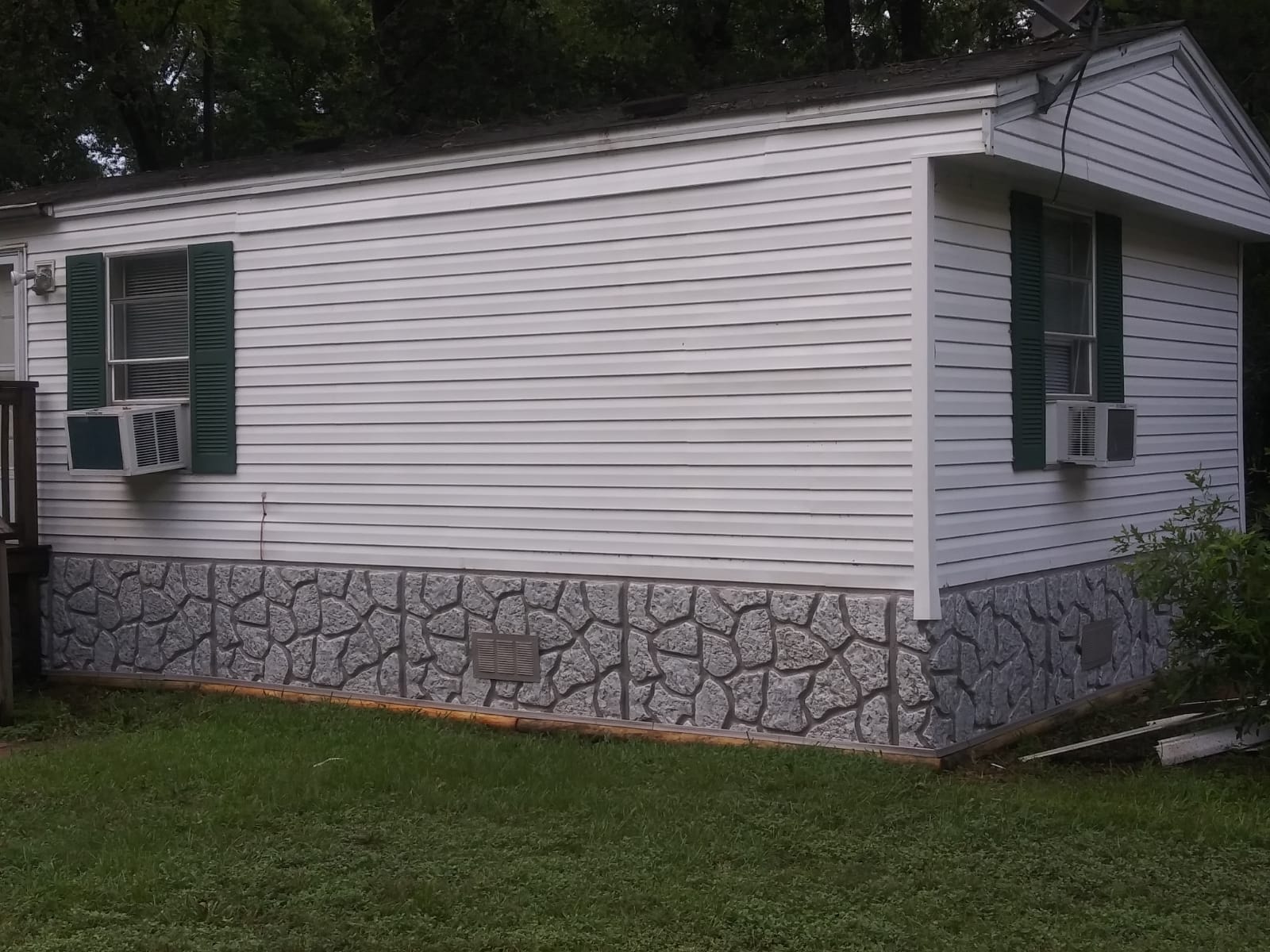 White mobile home with green shutters, window air conditioners, and stone-patterned skirting on a grassy lawn.