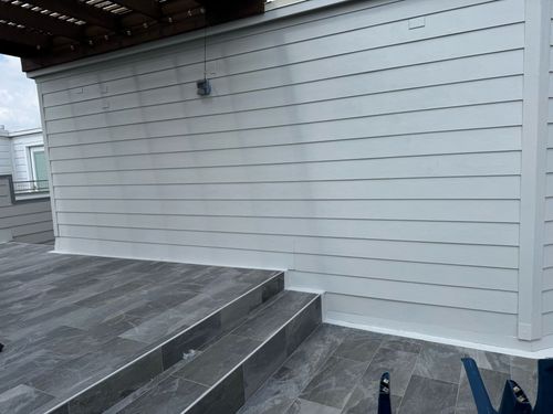 Gray tiled patio with a small set of stairs leading to a gray horizontal siding wall under a dark wood pergola.