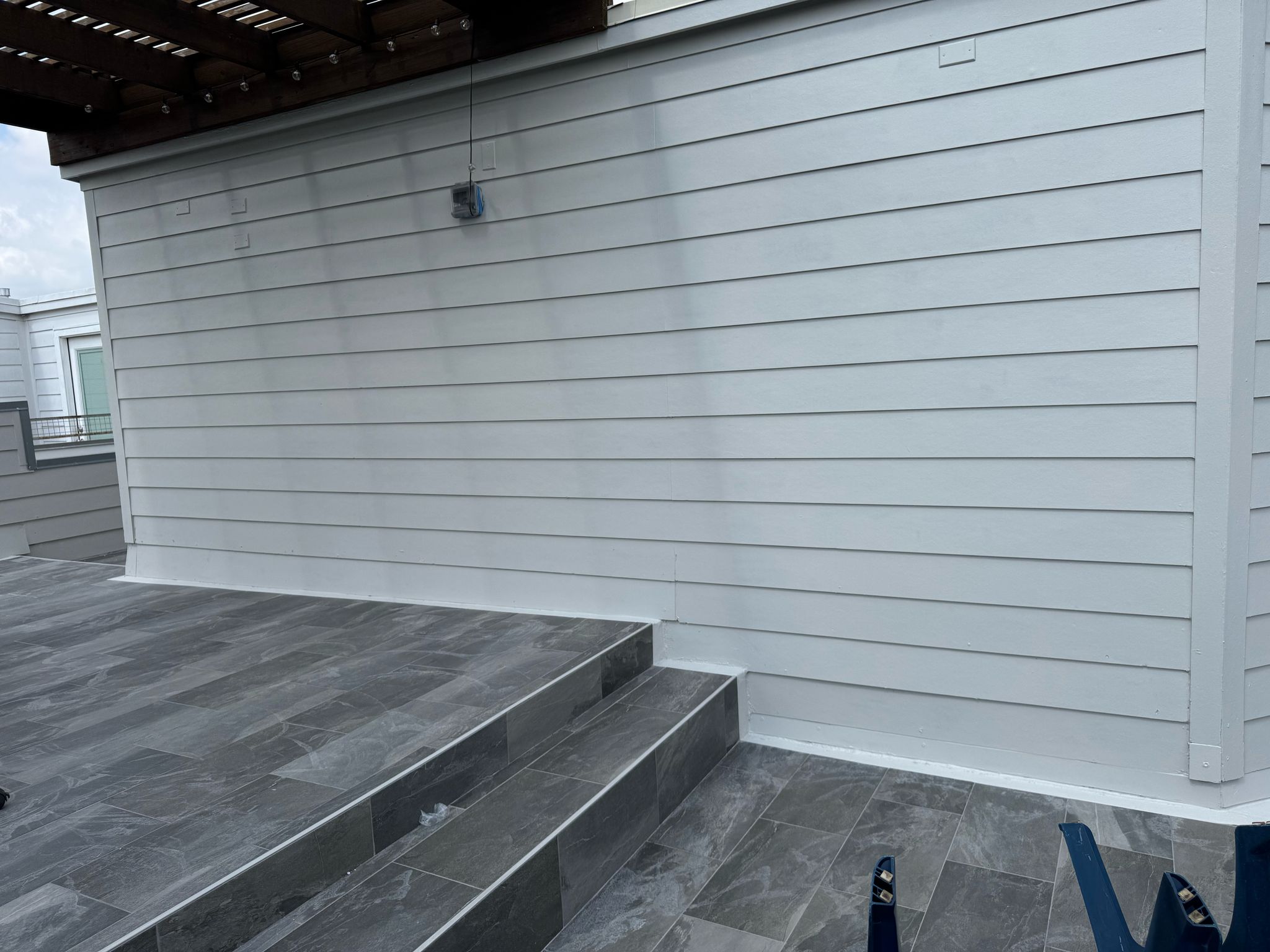 Gray tiled patio with a small set of stairs leading to a gray horizontal siding wall under a dark wood pergola.
