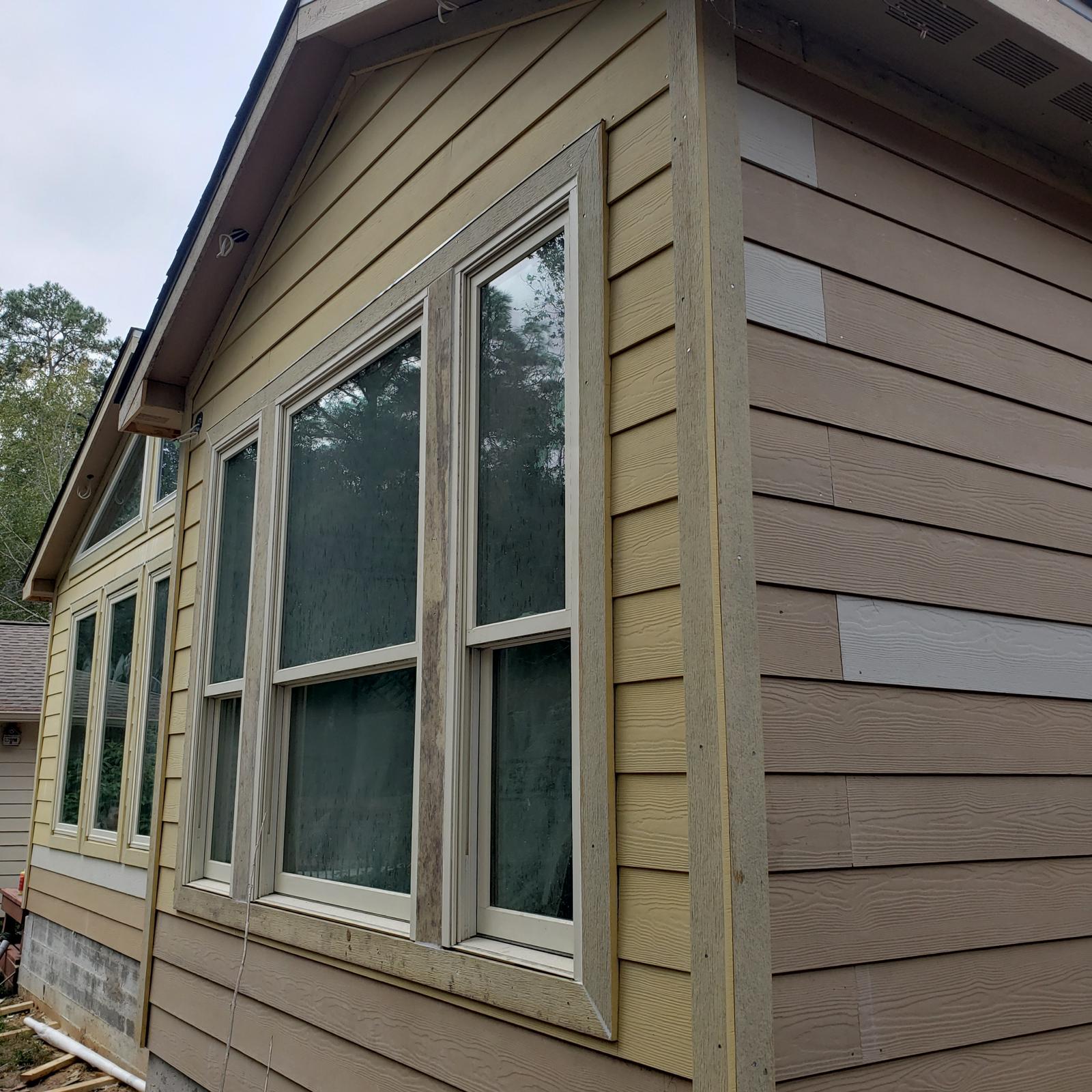 Exterior corner of a house showing tan and brown wood siding and multiple large windows with white frames reflecting trees.