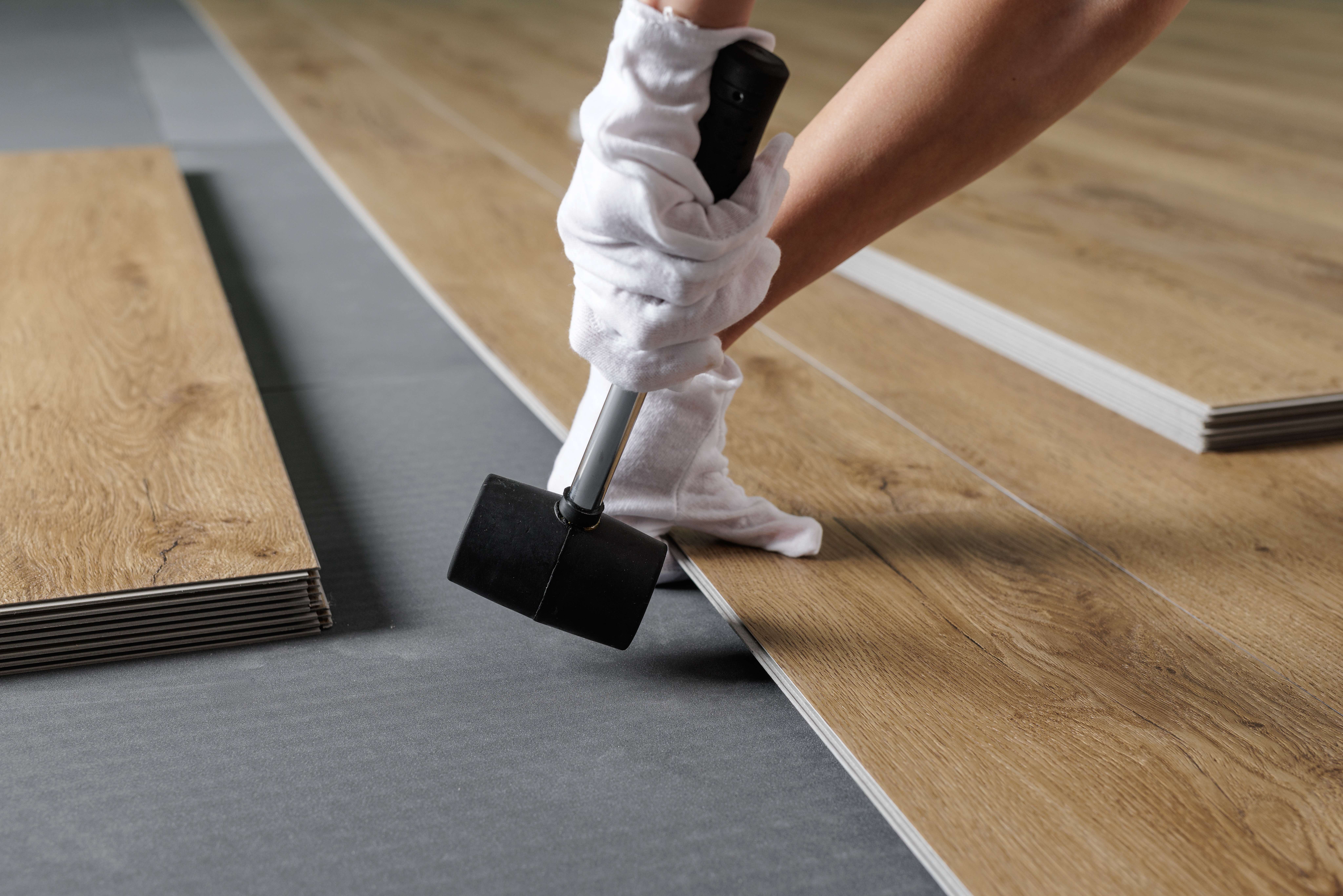 Person wearing white gloves installing wood-patterned vinyl plank flooring with a black rubber mallet.