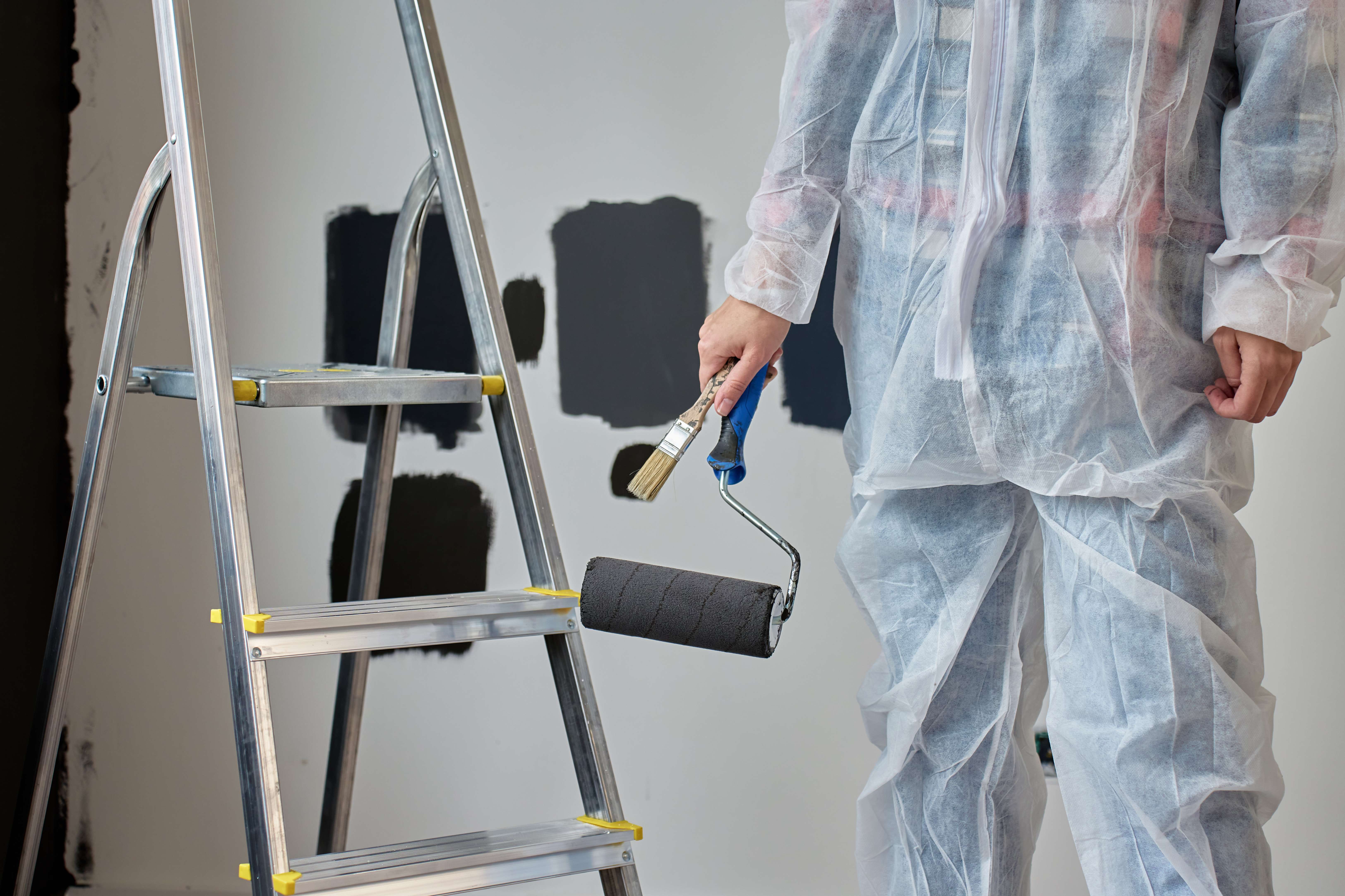 Person in protective coveralls holding a paint roller and brush near a metal stepladder with dark paint swatches on the wall.
