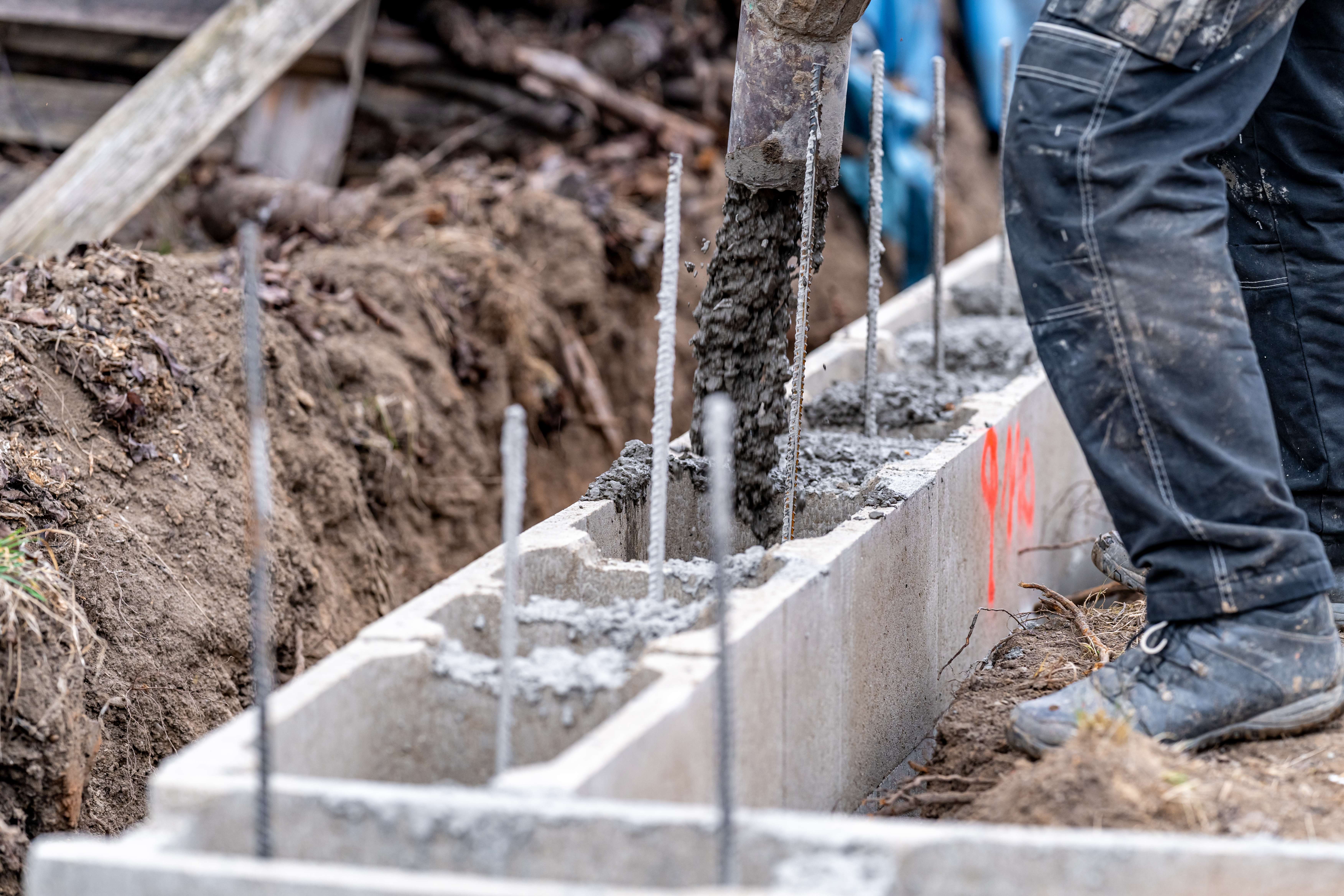 Construction worker pouring concrete into a cinder block foundation with exposed rebar rods.