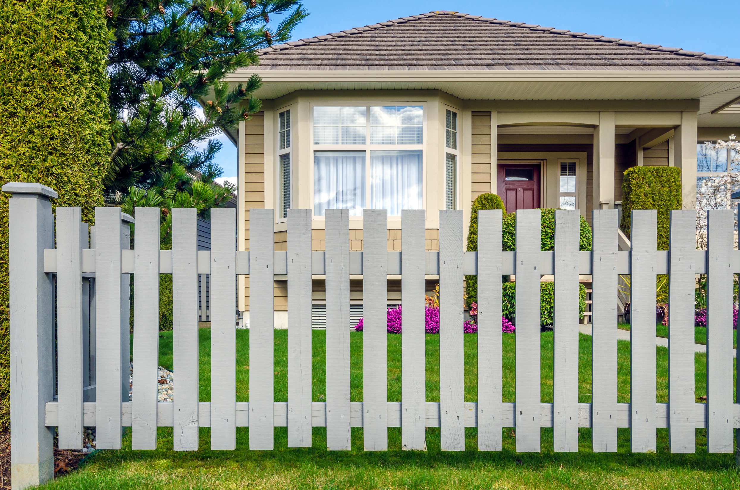 Front view of a beige house with a brown shingled roof behind a white picket fence and green lawn.