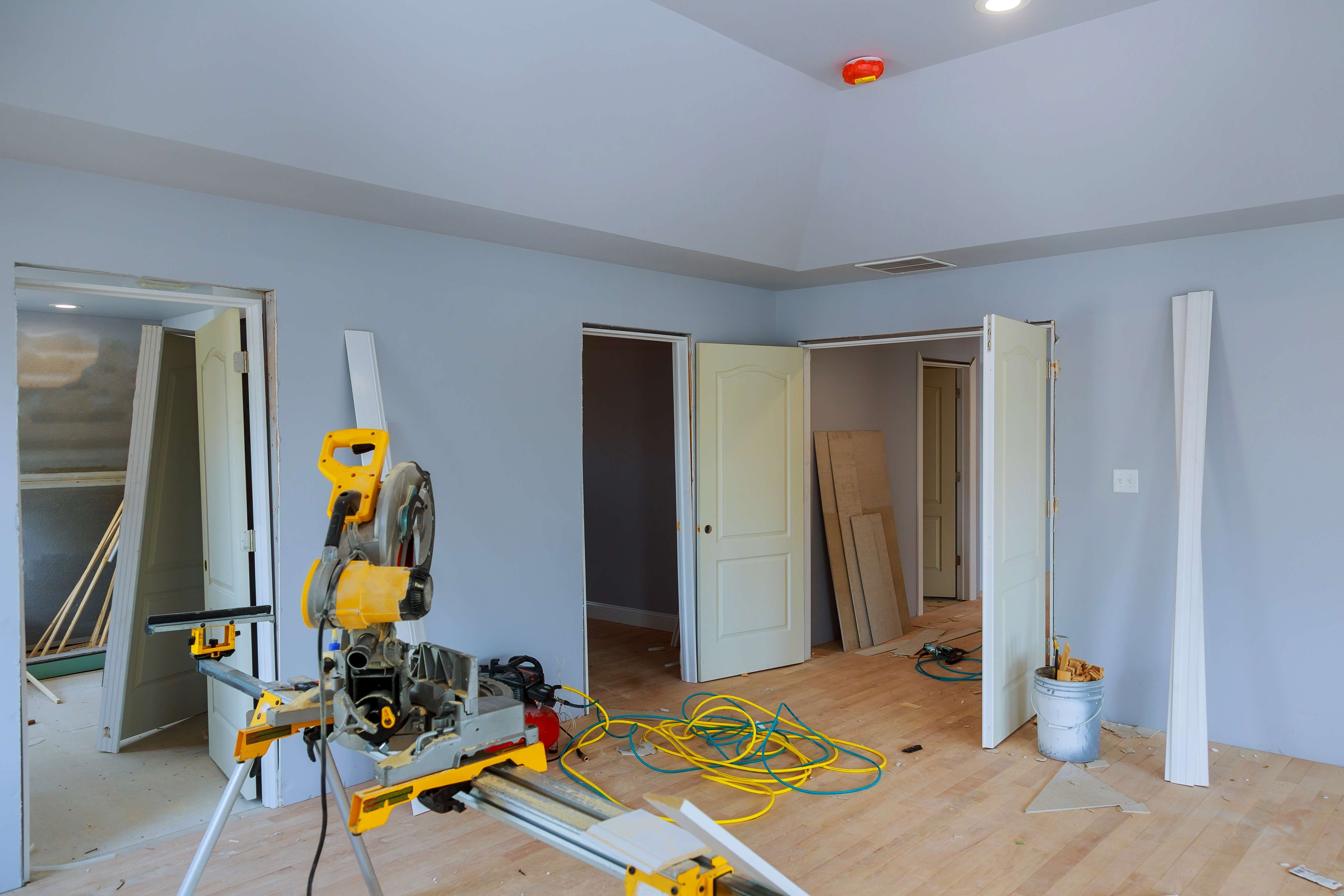Interior room under renovation with light wood flooring, open doors, a yellow miter saw on a stand, and scattered construction materials and cables.