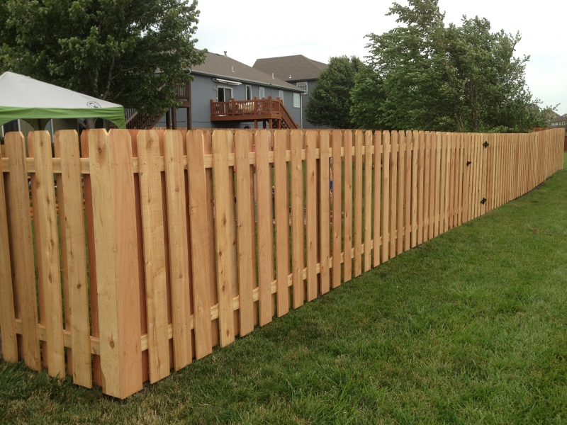 Long wooden picket fence running alongside a grassy yard with houses and trees in the background.