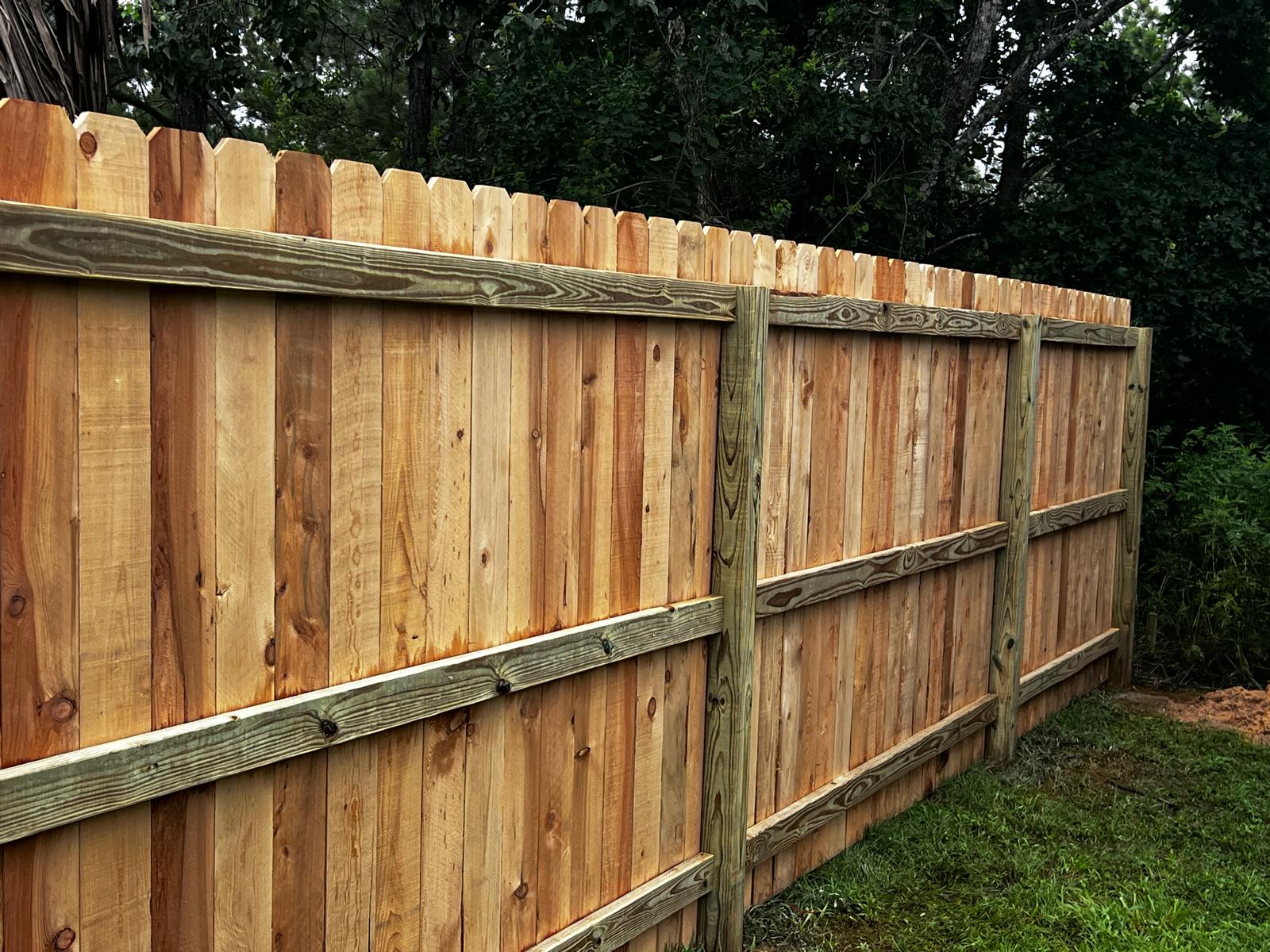 Newly installed wooden privacy fence with vertical planks and horizontal support beams beside grass and trees.