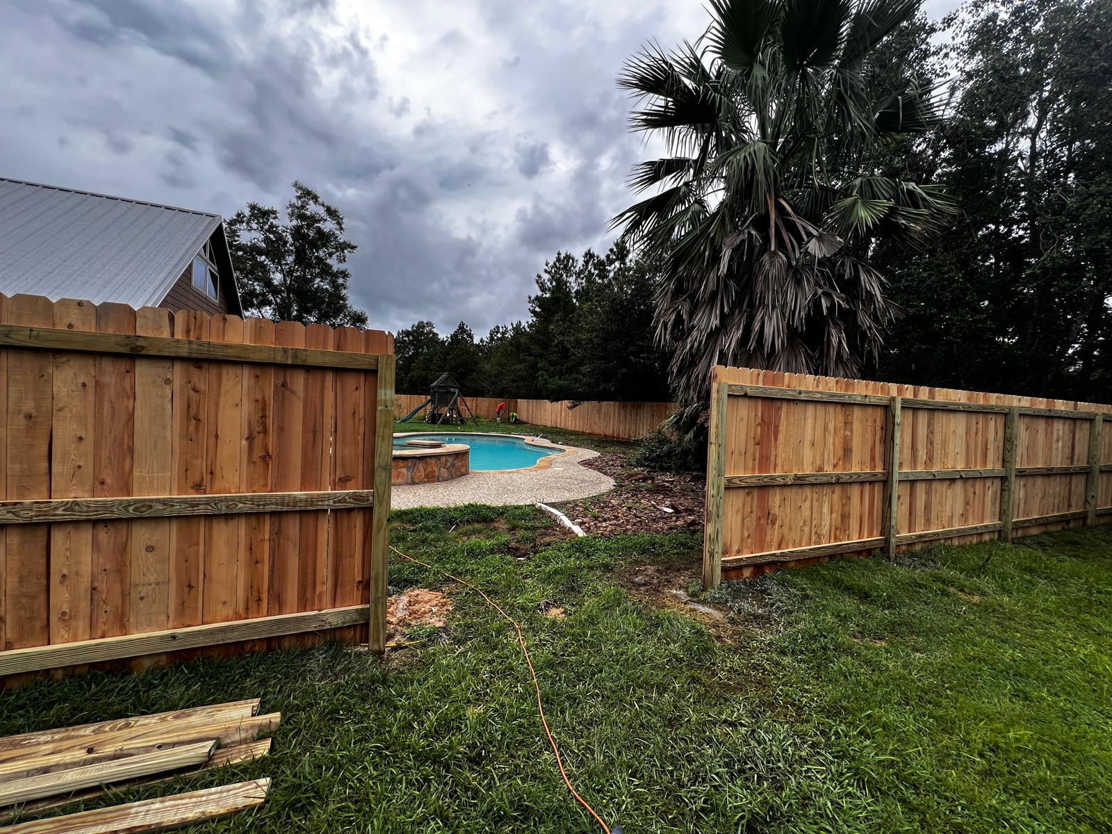 Newly installed wooden fence panels with an open gate leading to a backyard with a swimming pool and palm trees under a cloudy sky.
