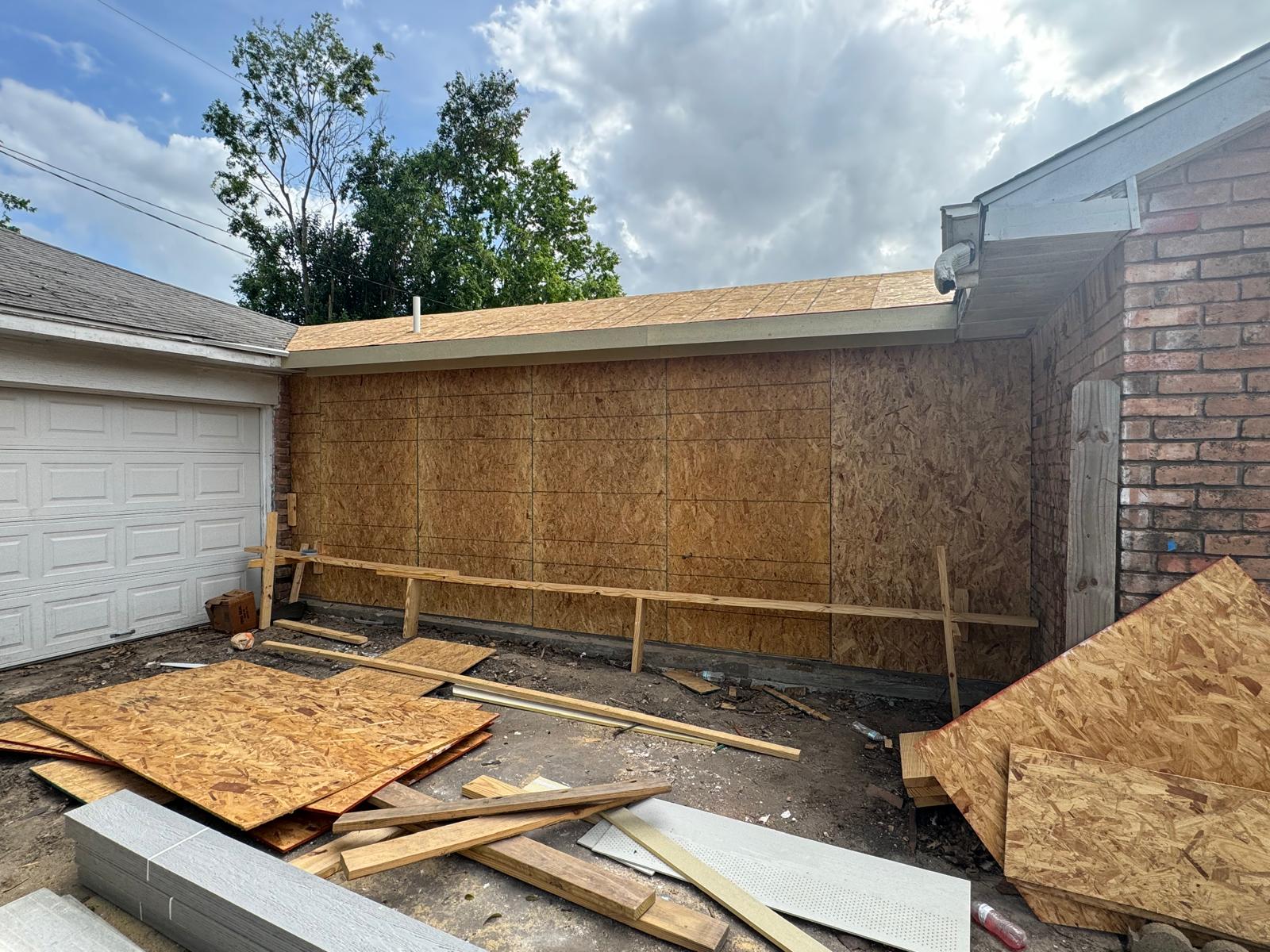 Construction site showing a partially built wooden exterior wall next to a white garage door, with scattered wood panels and building materials on the ground under a cloudy sky.