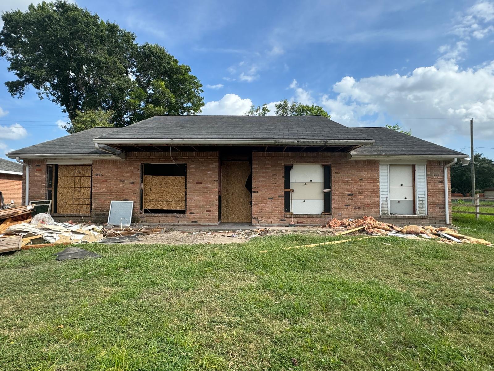 Single-story brick house with boarded-up windows and door, surrounded by scattered debris and renovation materials on the lawn.