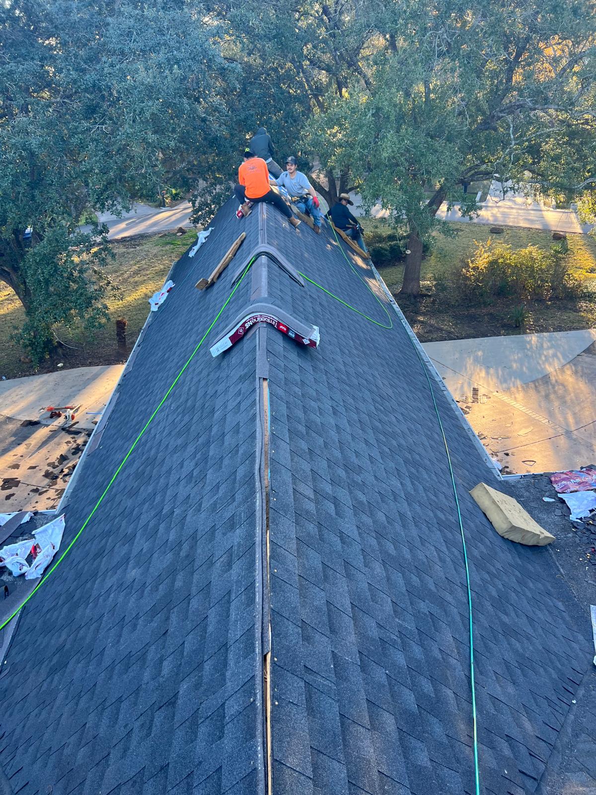 A group of workers installing dark gray asphalt shingles on a steep residential roof surrounded by trees and sunlight.