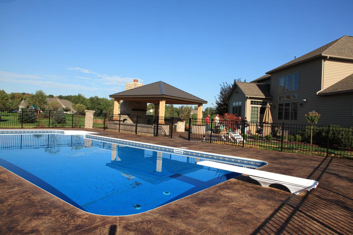 Backyard with a blue swimming pool, diving board, black metal fence, wooden gazebo, and a beige two-story house under a clear blue sky.