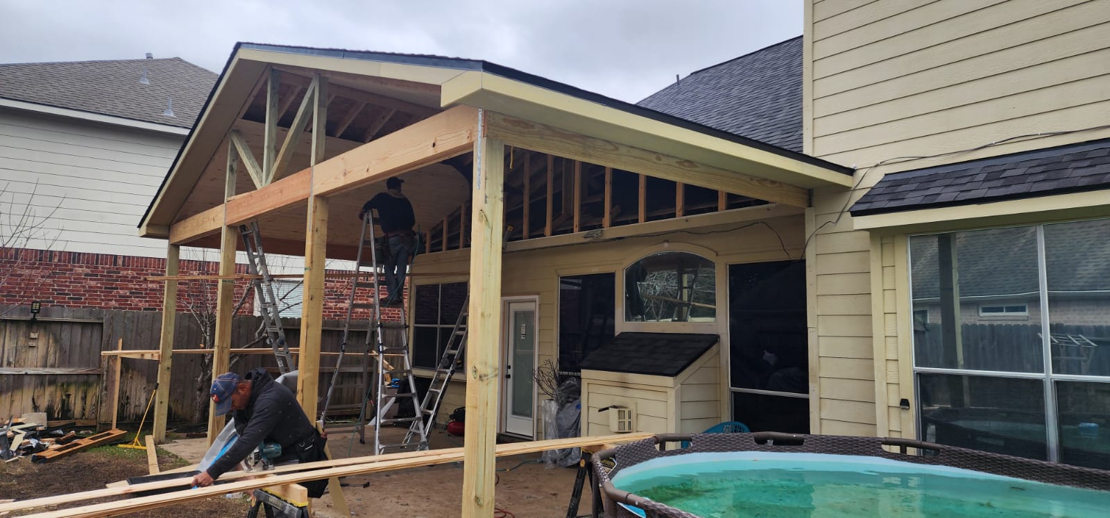 Two construction workers building a wooden covered patio attached to a house beside a small round pool.