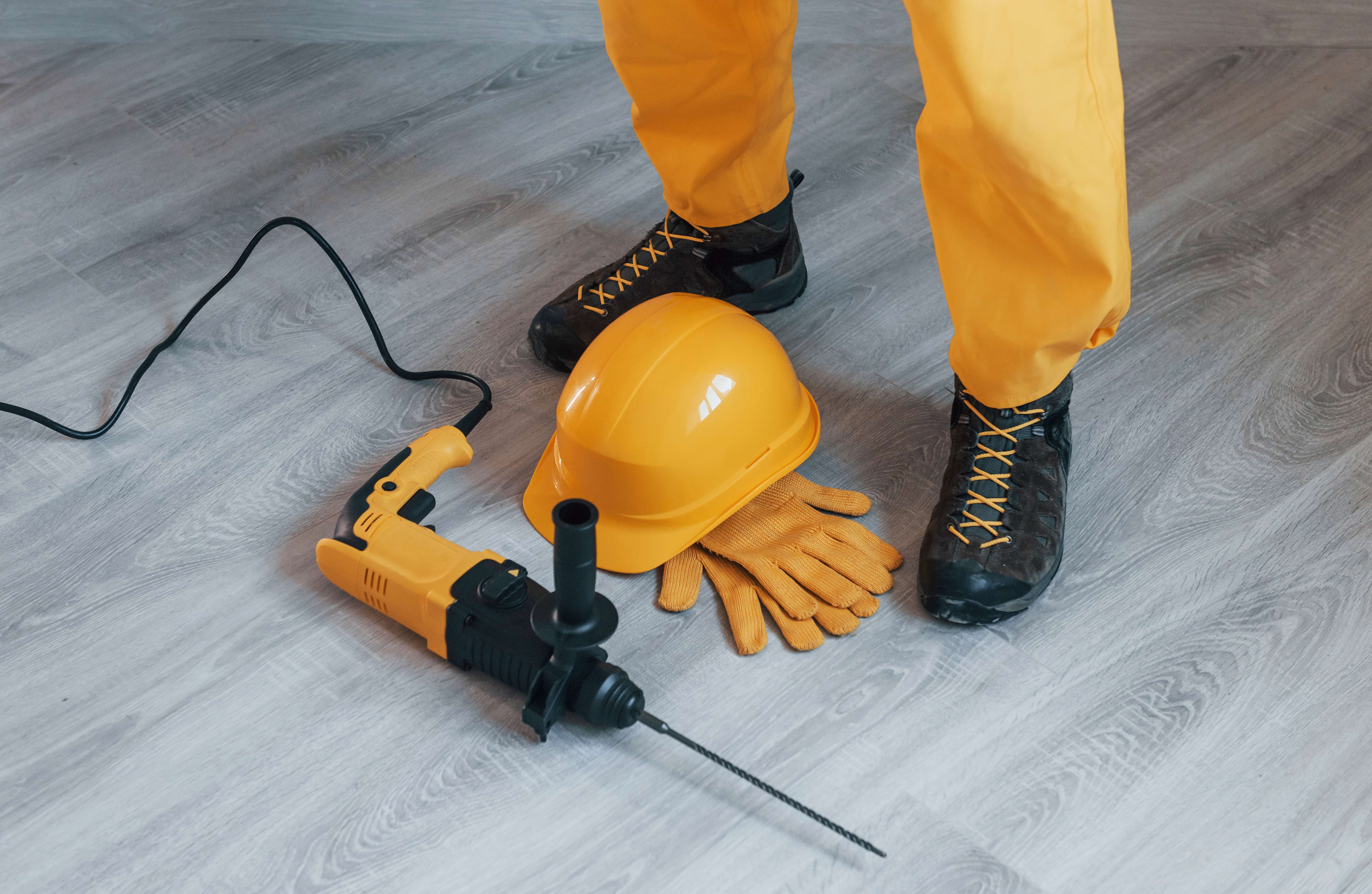 Person in yellow pants and black boots standing next to a yellow hard hat, yellow work gloves, and a yellow and black power drill on a gray wood floor.