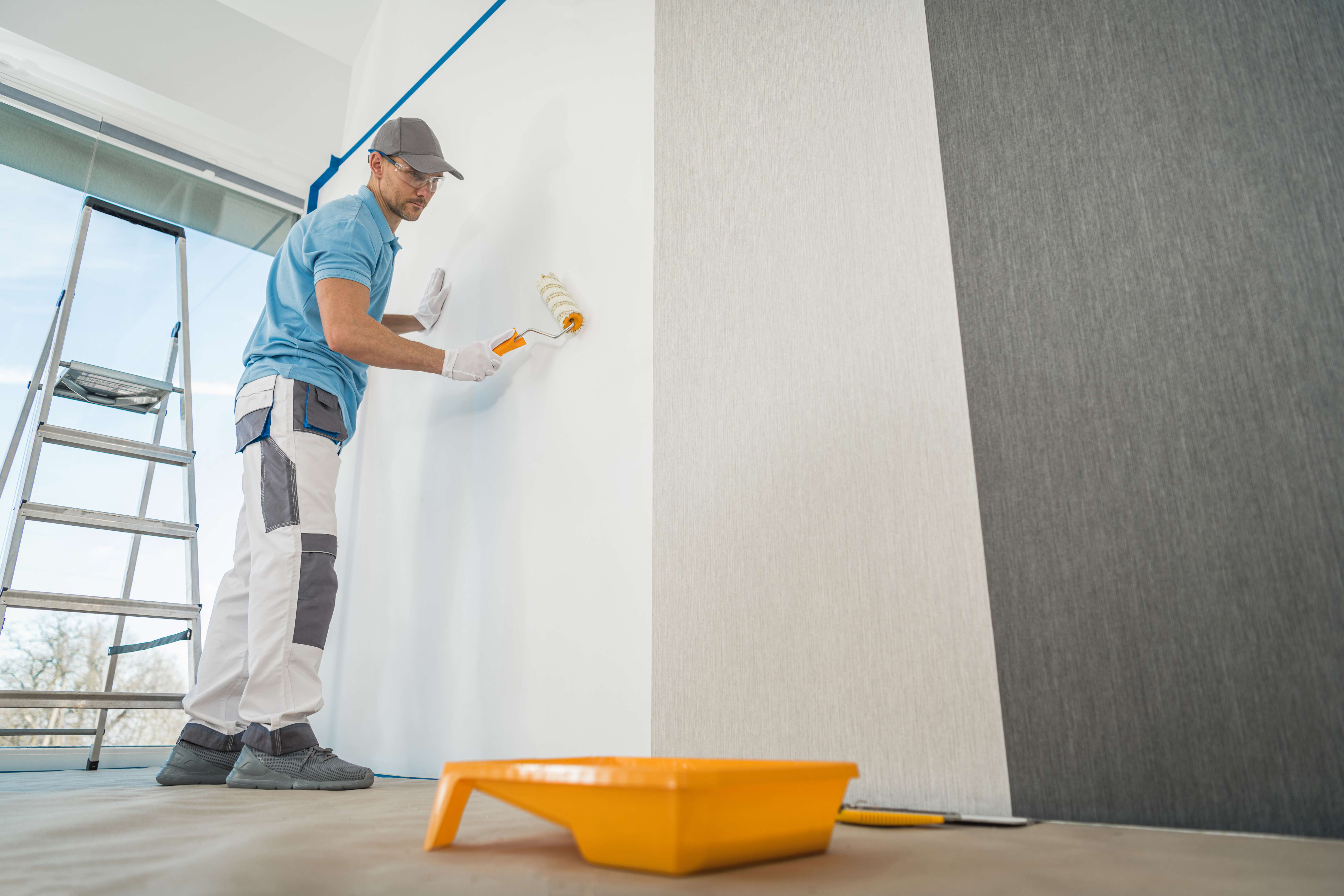 Man wearing gloves and cap using a paint roller to apply adhesive on a white wall next to wallpaper rolls.