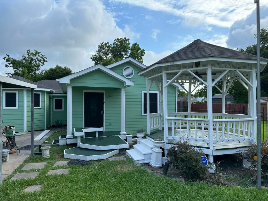 Green house with white trim and black door next to a white wooden gazebo in a grassy backyard under a partly cloudy sky.