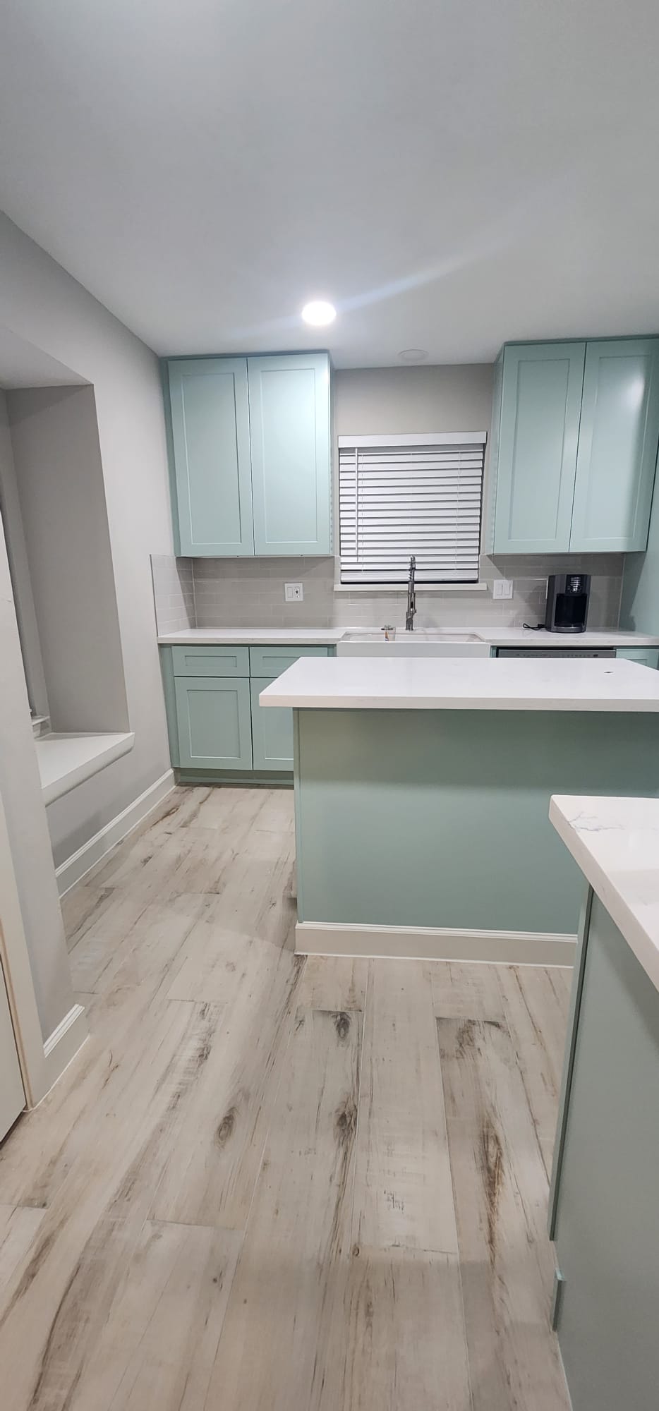 Modern kitchen with light blue cabinets, white countertops, farmhouse sink below a window with blinds, light wood flooring, and a built-in seating nook on the left.