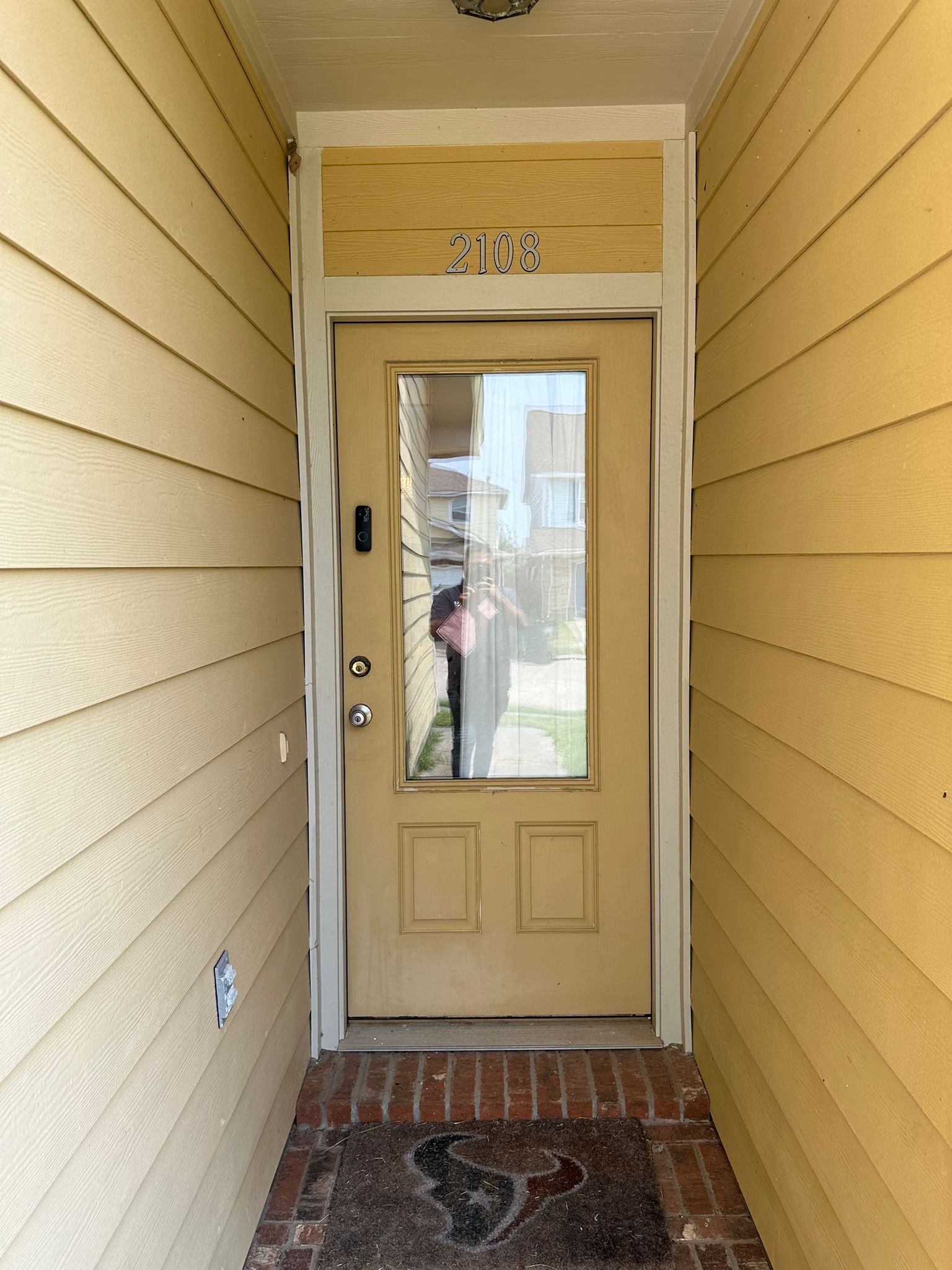 Yellow front door with a large central window and house number 2108 above, framed by yellow siding and a brick doorstep with a Houston Texans logo doormat.