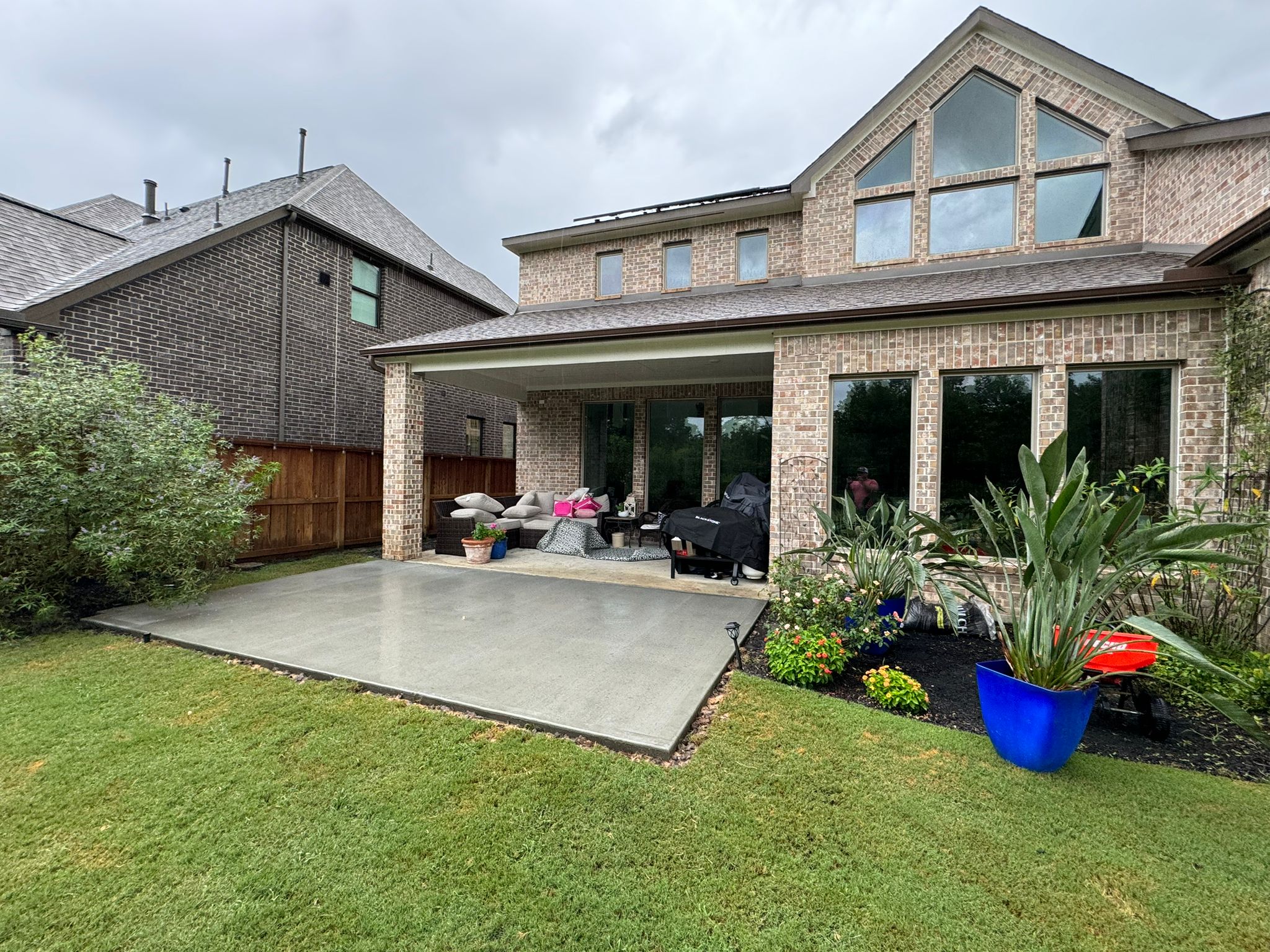 Backyard view of a brick house with a covered patio furnished with a sofa and grill, adjacent to a concrete slab and green lawn.