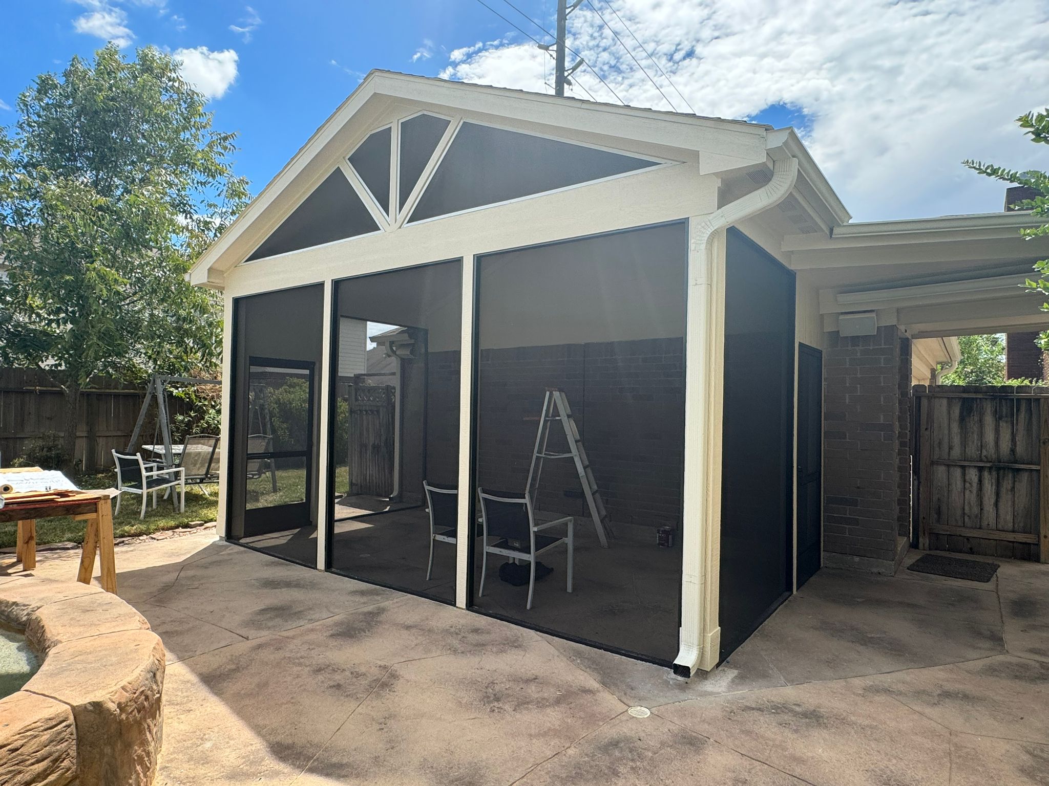 Screened patio enclosure with a pitched roof, two chairs, and a ladder inside, next to a stone-edged pool and backyard fence.