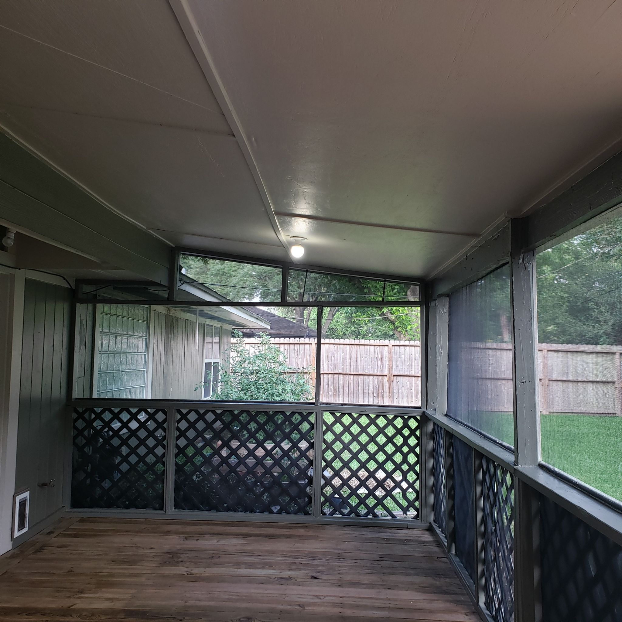 Empty screened-in porch with wooden floor, lattice panels, and a light bulb on the ceiling, overlooking a fenced backyard.