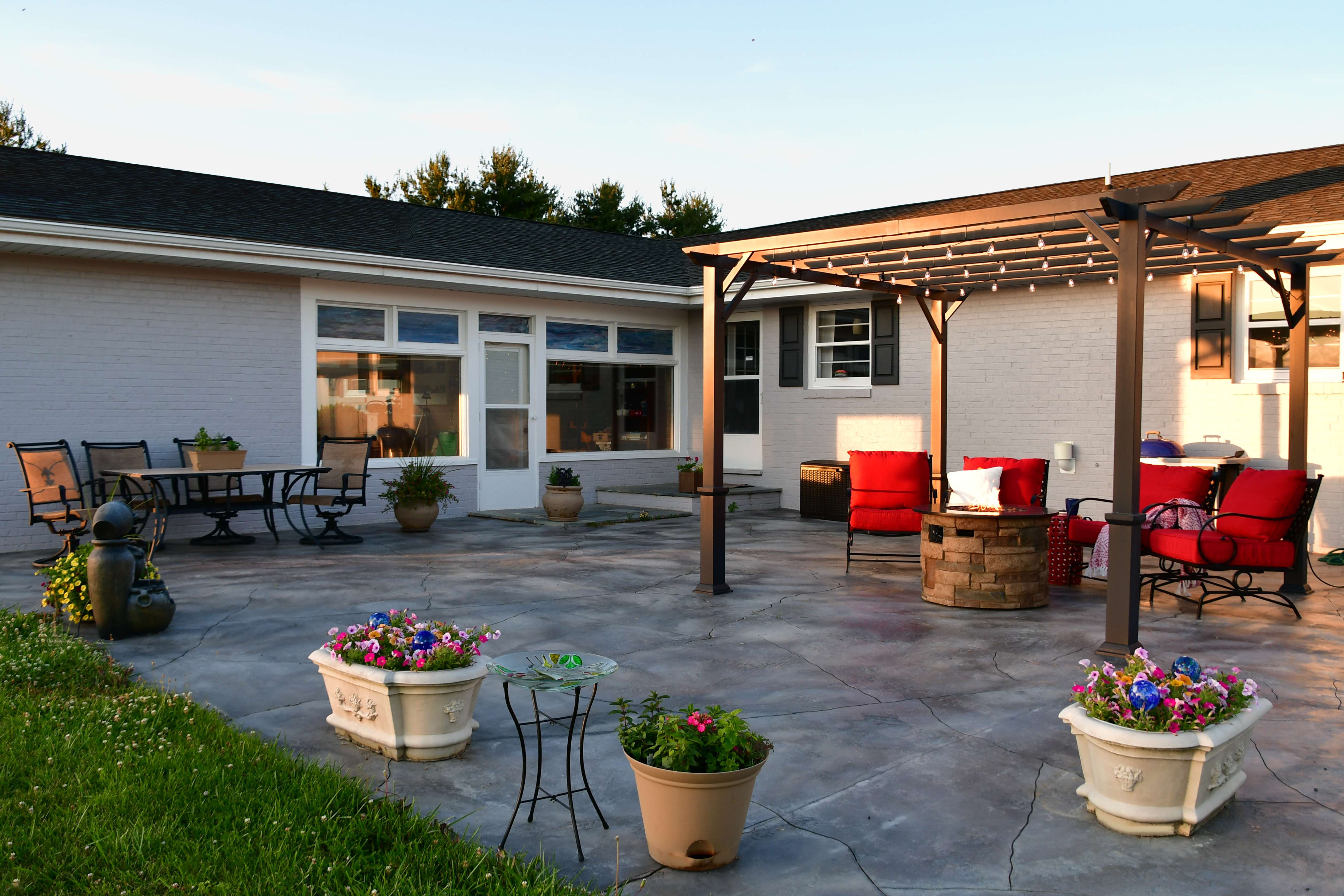 Patio with concrete floor featuring a seating area under a pergola with string lights, red cushioned chairs around a fire pit, flower pots, and a dining set near a white brick house.