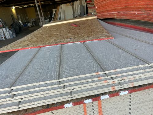 Stack of gray wooden siding panels with grooved texture and a piece of plywood on top in a warehouse.