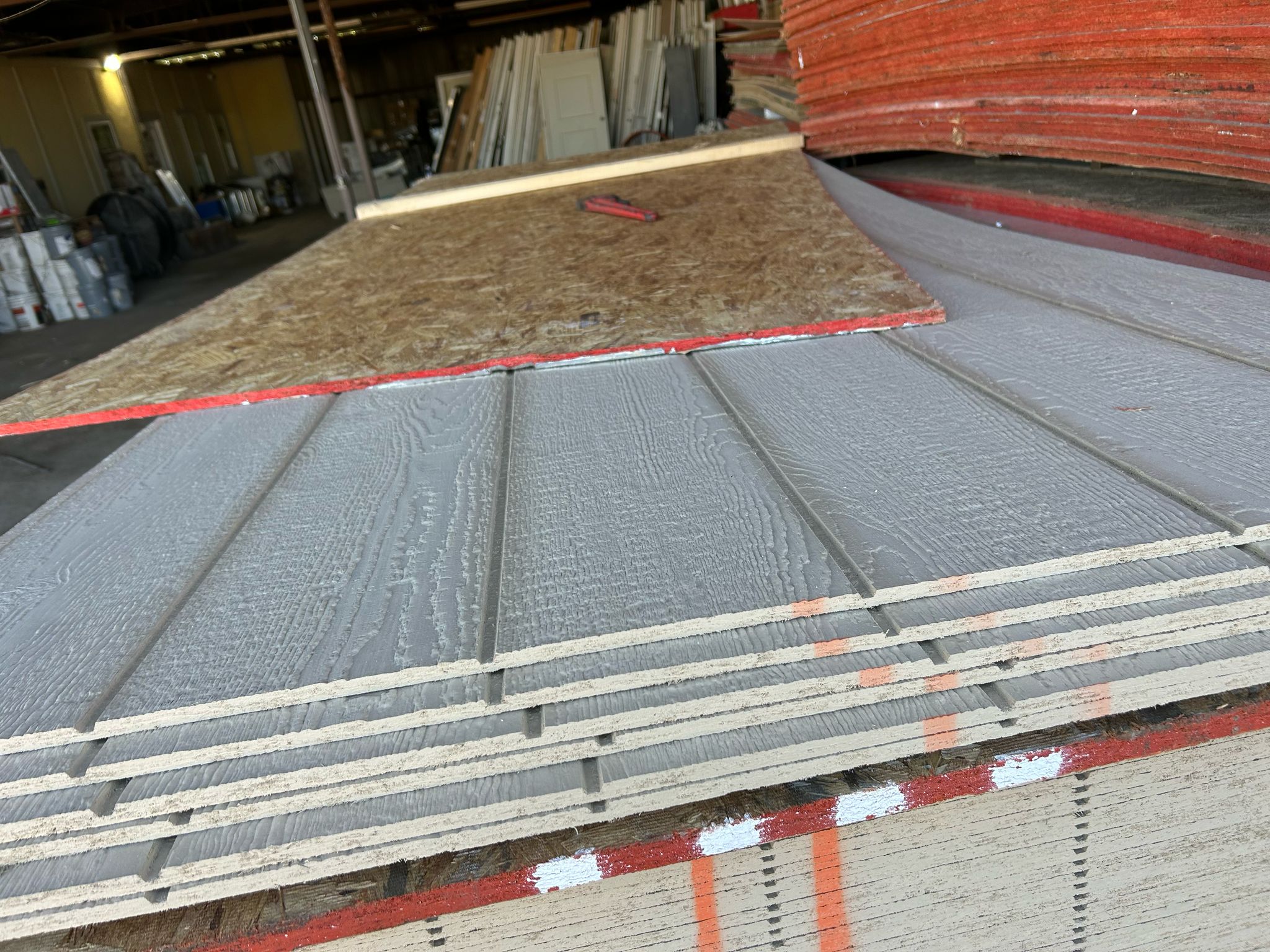 Stack of gray wooden siding panels with grooved texture and a piece of plywood on top in a warehouse.