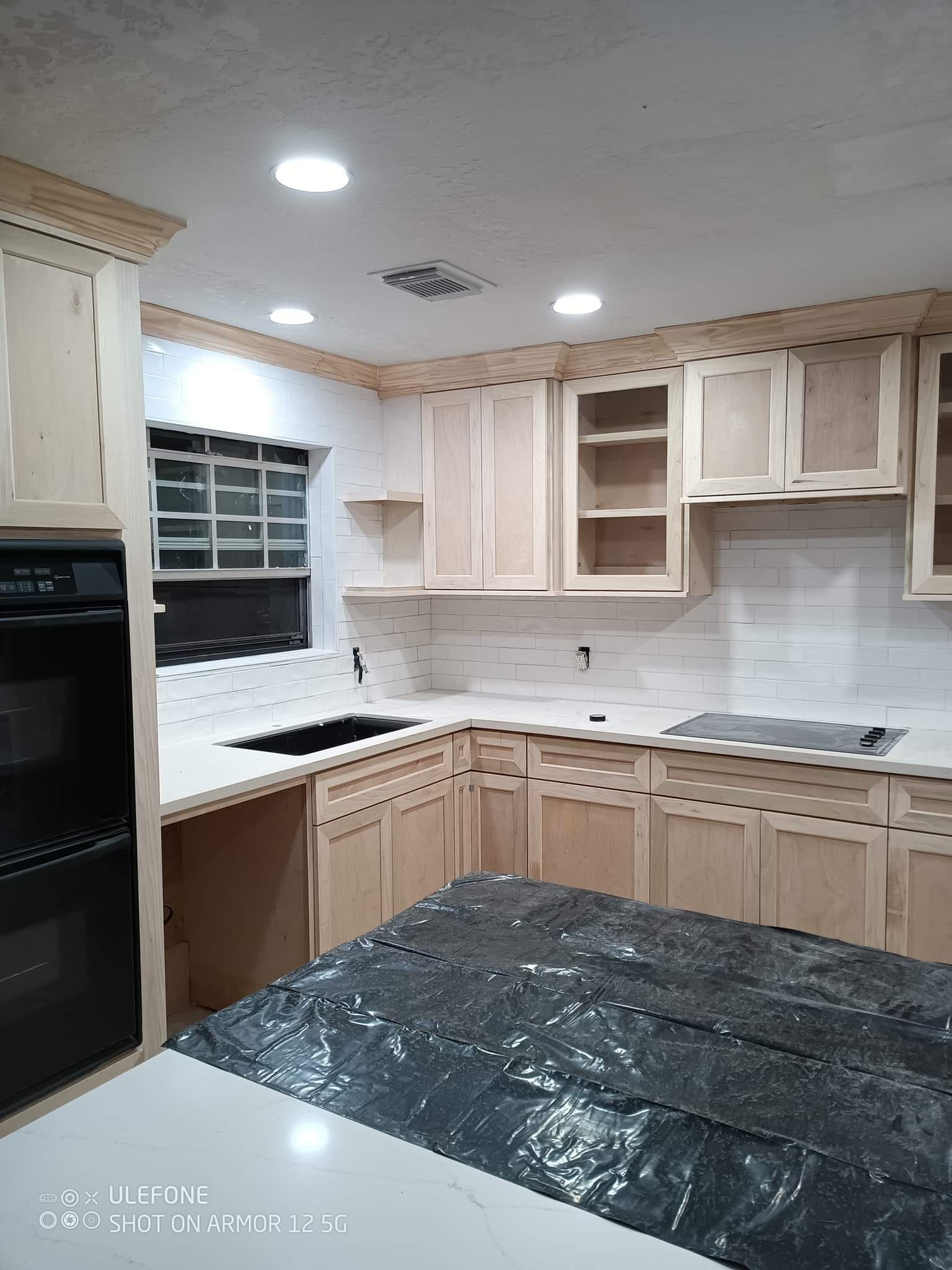 Modern kitchen with light wood cabinets, white countertops, black sink, and built-in stovetop, under recessed ceiling lights.