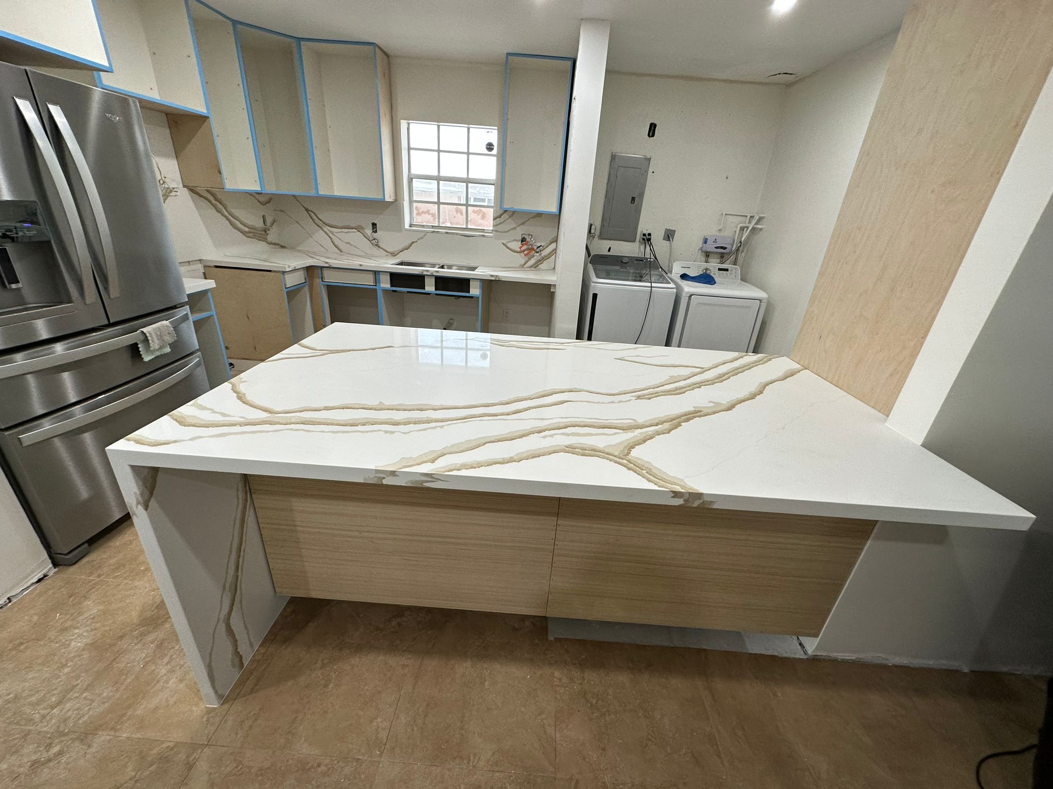 Modern kitchen under renovation with marble-patterned countertops and island, light wooden cabinets, stainless steel refrigerator, and laundry area with washer and dryer.