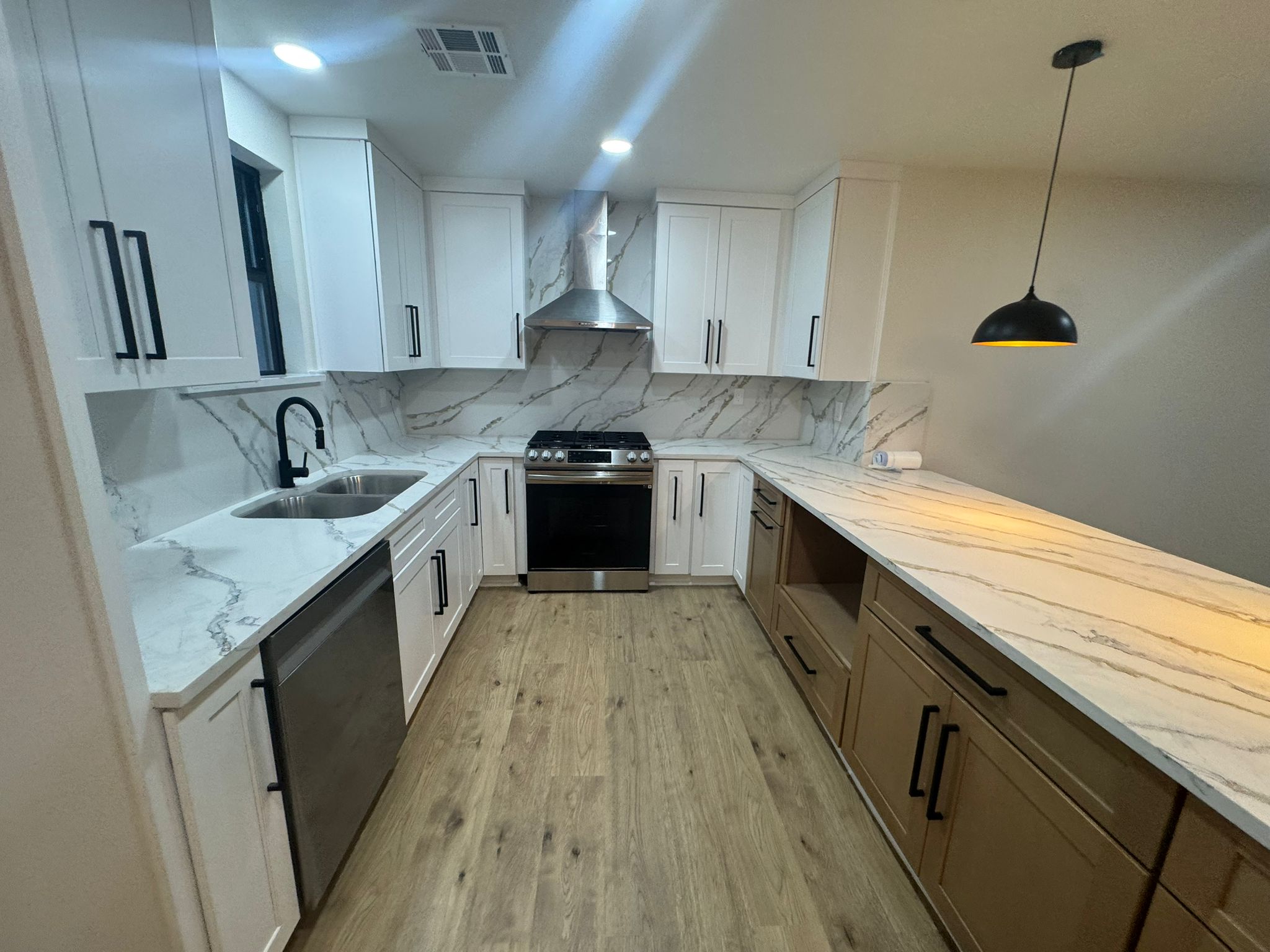 Modern kitchen with white upper cabinets, brown lower cabinets, marble-patterned countertops and backsplash, stainless steel appliances, wood floor, and a black pendant light.