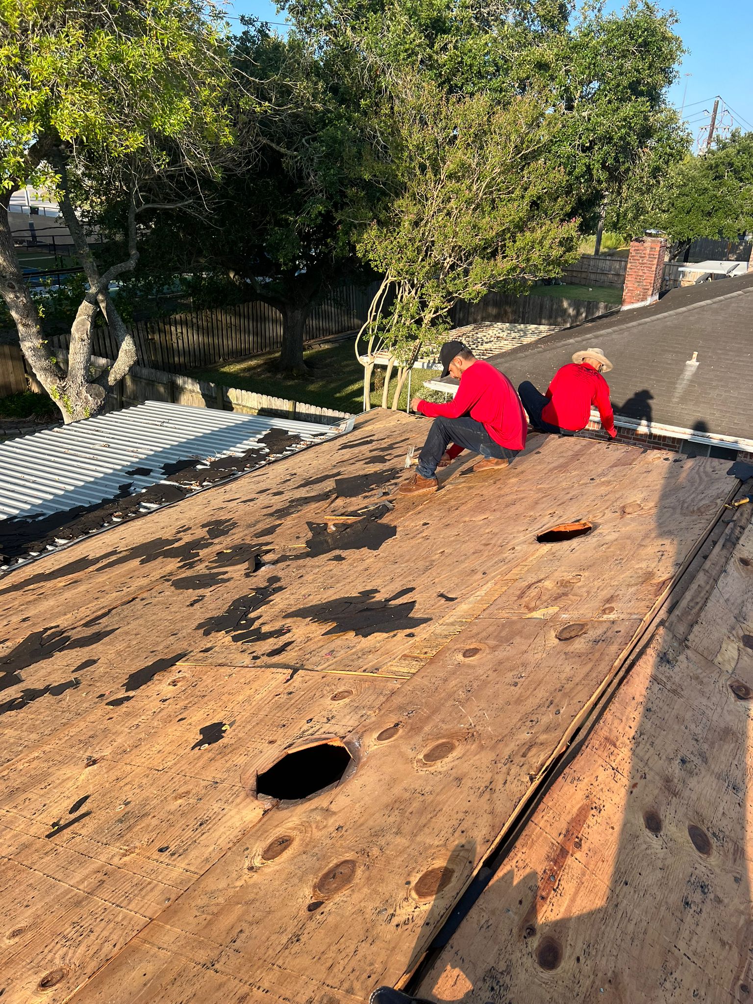 Two roofers in red shirts working on a wooden roof deck with damaged holes and peeling black material.