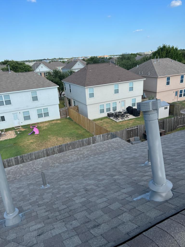 View of a gray shingled rooftop with vent pipes, overlooking fenced backyards and neighboring two-story houses under a clear blue sky.